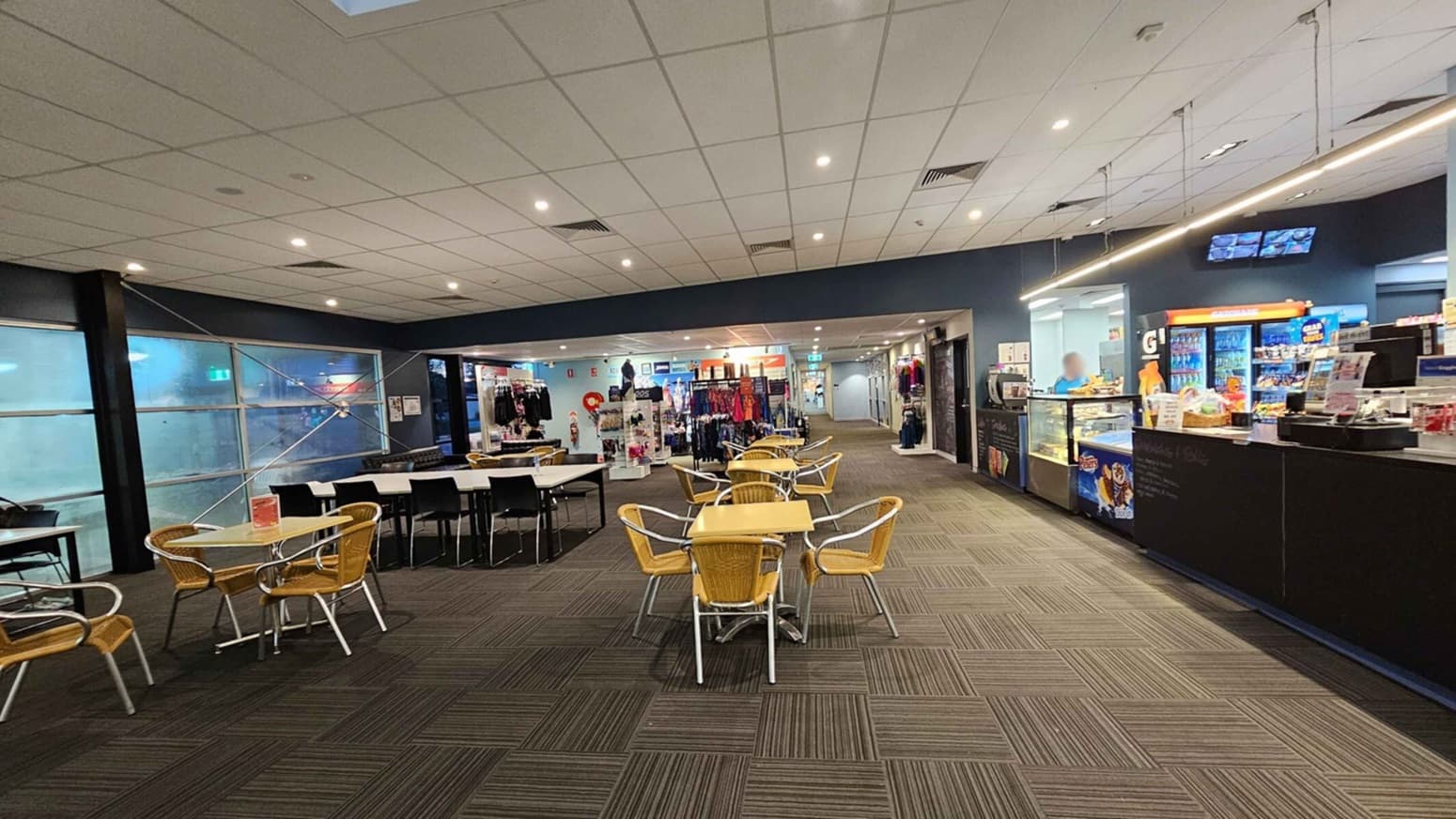 A spacious indoor area with a café on the right, featuring a counter displaying snacks and drinks. A person is behind the counter. In the centre, there are tables and chairs with wicker seats. On the left, a row of tables with black chairs is positioned near large frosted windows. In the background, a shop displays clothing and accessories. The ceiling is fitted with recessed lighting, and the floor is covered with patterned carpet tiles.