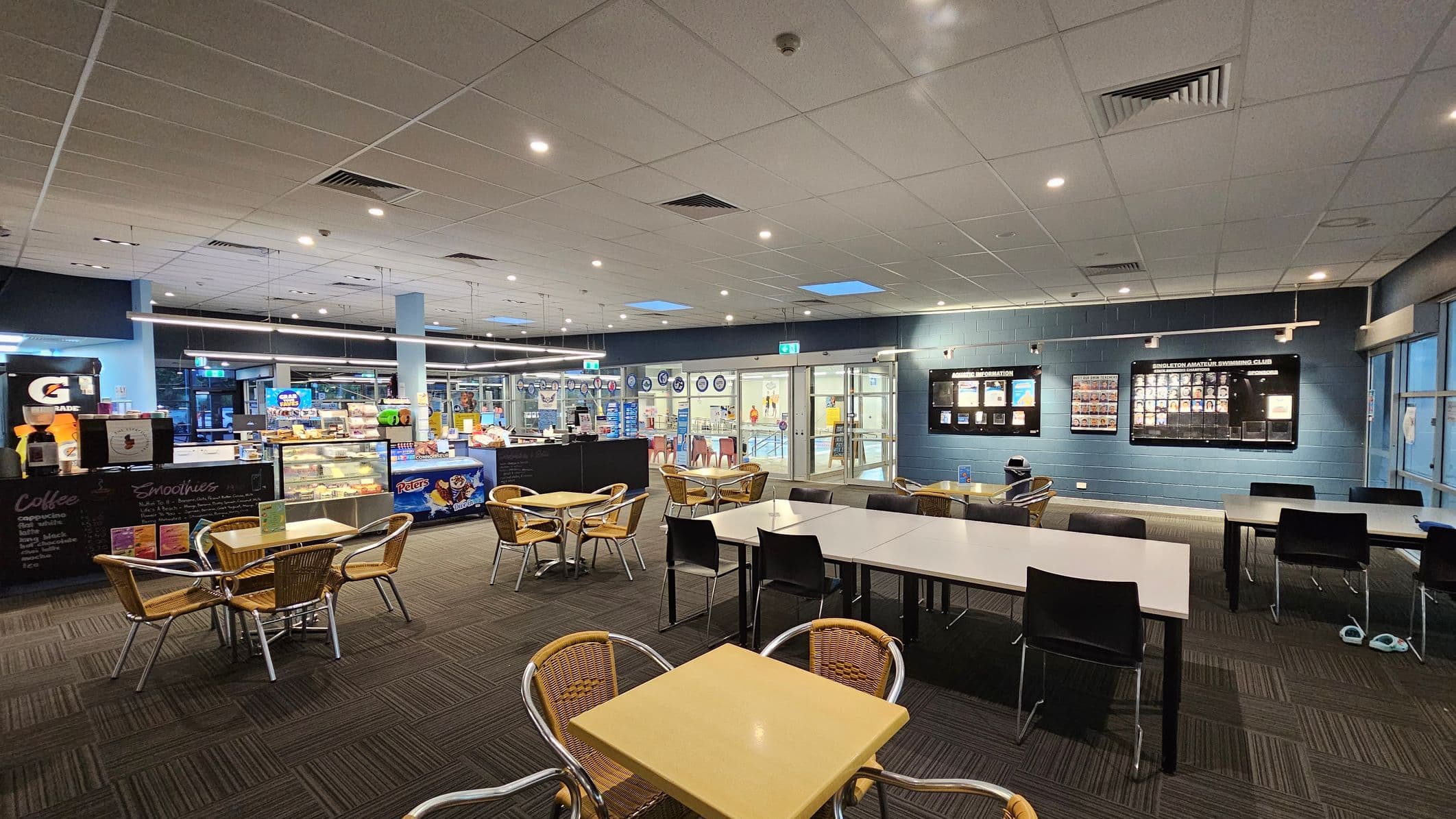 A spacious indoor café area with a tiled ceiling and carpeted floor. On the left, there is a counter displaying snacks and drinks, with a Gatorade dispenser and a menu board for coffee and smoothies. In the centre, several tables with wicker chairs are arranged. On the right, a wall features display boards with information and photos. The background shows glass doors leading to another area, possibly a pool or gym.