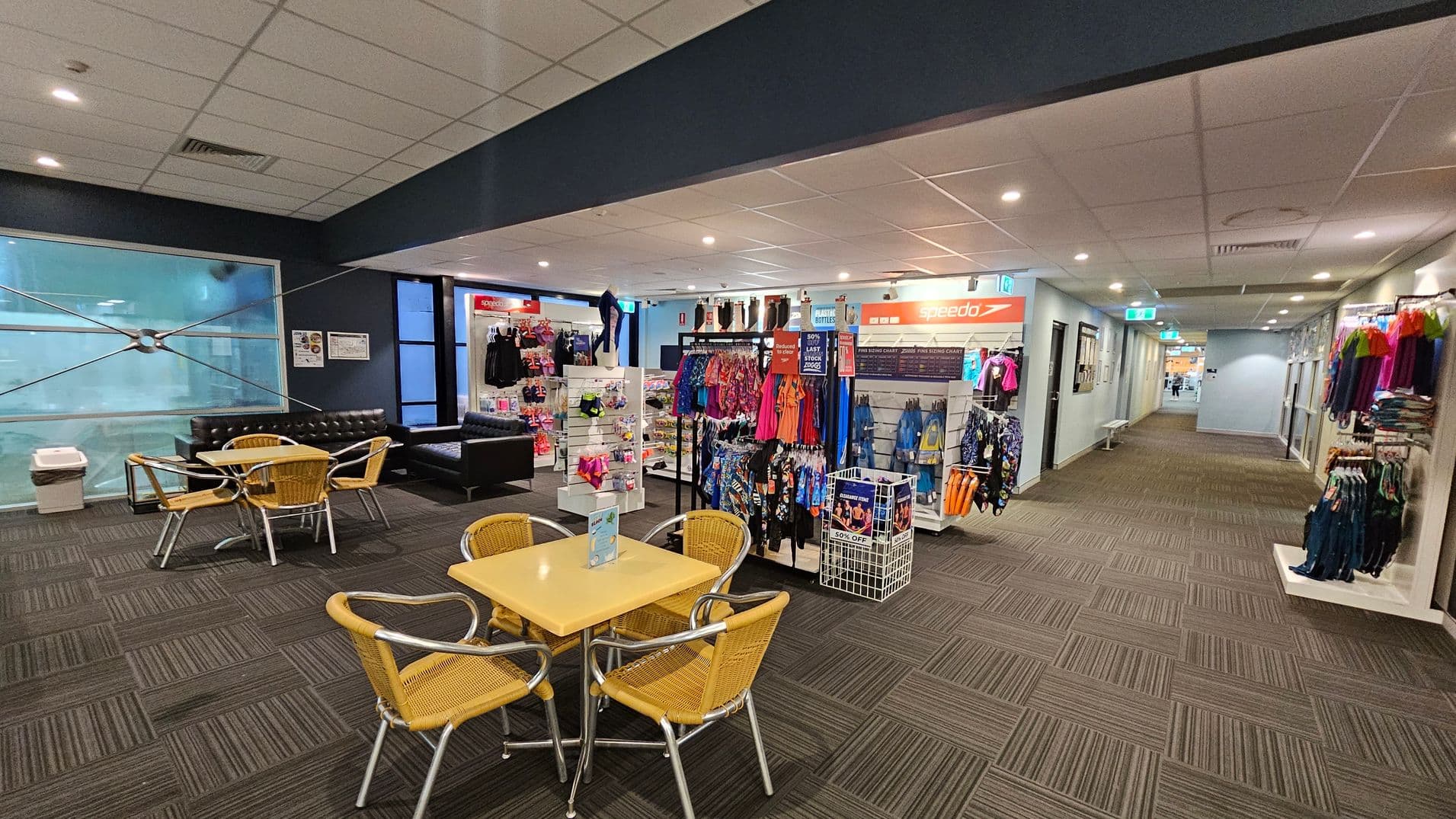 A retail area with a seating section on the left, featuring a yellow table and wicker chairs. Behind the seating, there are black leather sofas and a large window. In the centre, there are displays of swimwear and accessories, with racks of colourful clothing and a Speedo sign above. On the right, a corridor leads further into the building, with more clothing racks along the wall. The floor is carpeted with a striped pattern, and the ceiling has recessed lighting.
