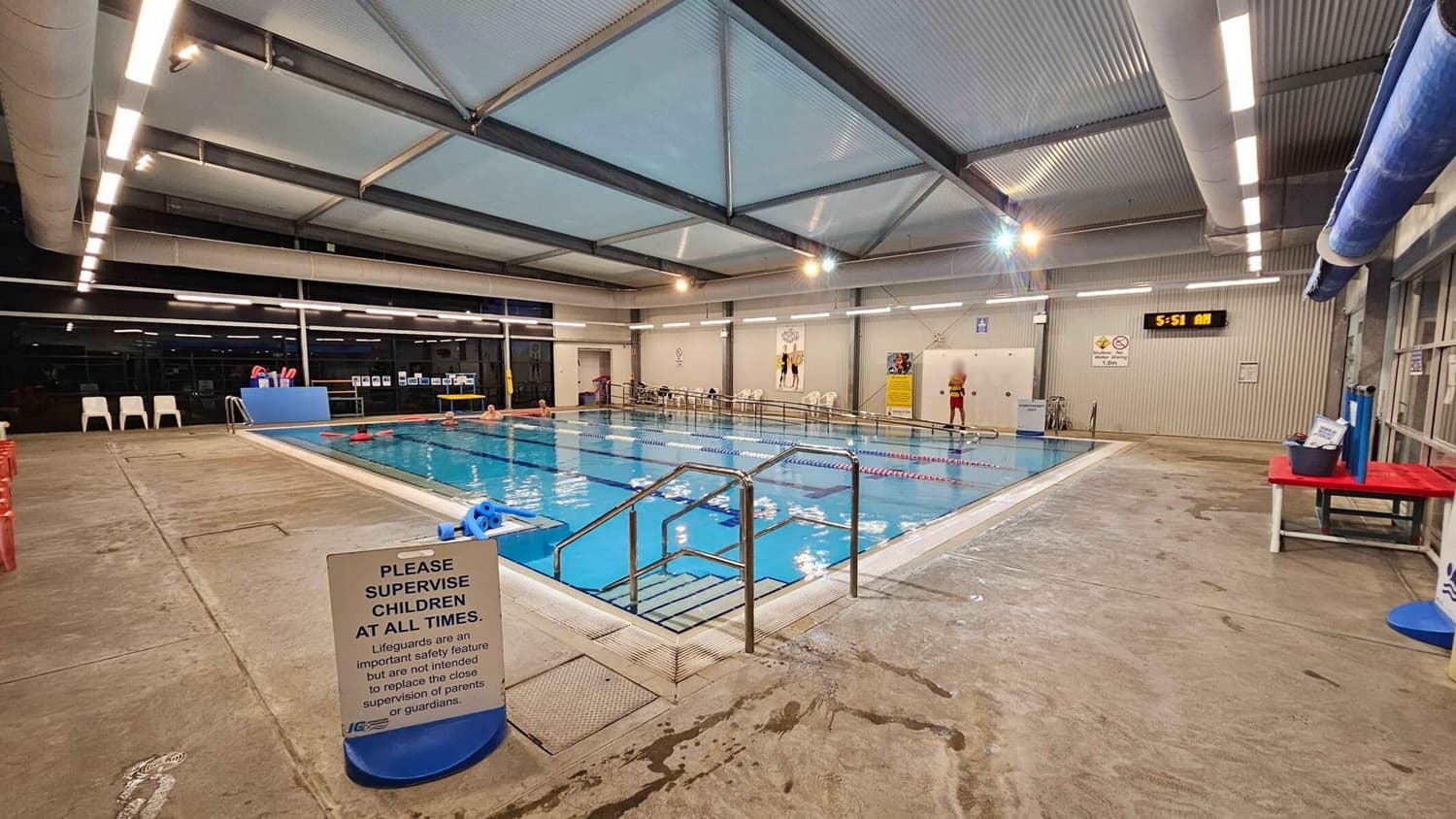 Indoor swimming pool with clear blue water, surrounded by a concrete floor. On the left, there are white plastic chairs and a blue sign reminding to supervise children. In the middle, a metal handrail leads into the pool. On the right, a lifeguard stands near a digital clock showing 5:51 AM. The ceiling has exposed beams and fluorescent lights. A red table with pool equipment is also visible on the right.