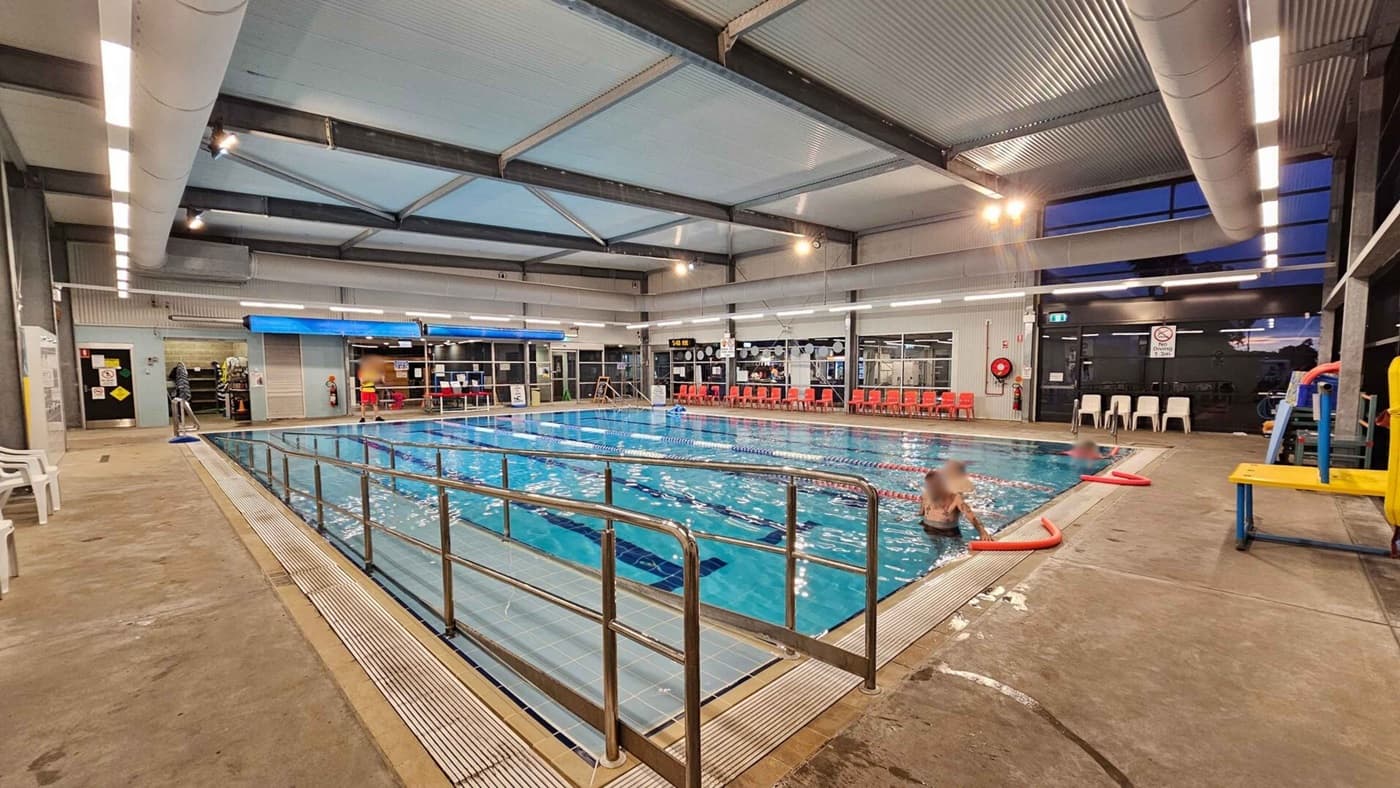 Indoor swimming pool with clear water, surrounded by a metal railing. On the left, there are white plastic chairs and a concrete floor. The right side has a few people in the pool and a yellow platform. The background features a wall with large windows, red chairs, and a lifeguard station. The ceiling is high with exposed beams and lighting fixtures.