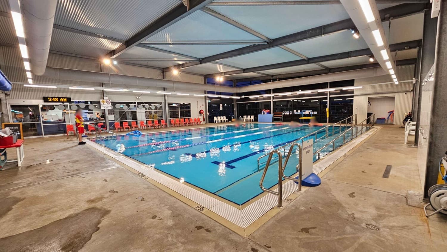 Indoor swimming pool with clear blue water, divided into lanes by floating ropes. On the left, a lifeguard in red and yellow stands by the poolside. Behind them, a digital clock displays 5:48 AM above a row of red chairs. On the right, a metal handrail leads into the pool, with a sign indicating "Hydrotherapy Only." The floor is wet concrete, and the ceiling has exposed beams and lighting.
