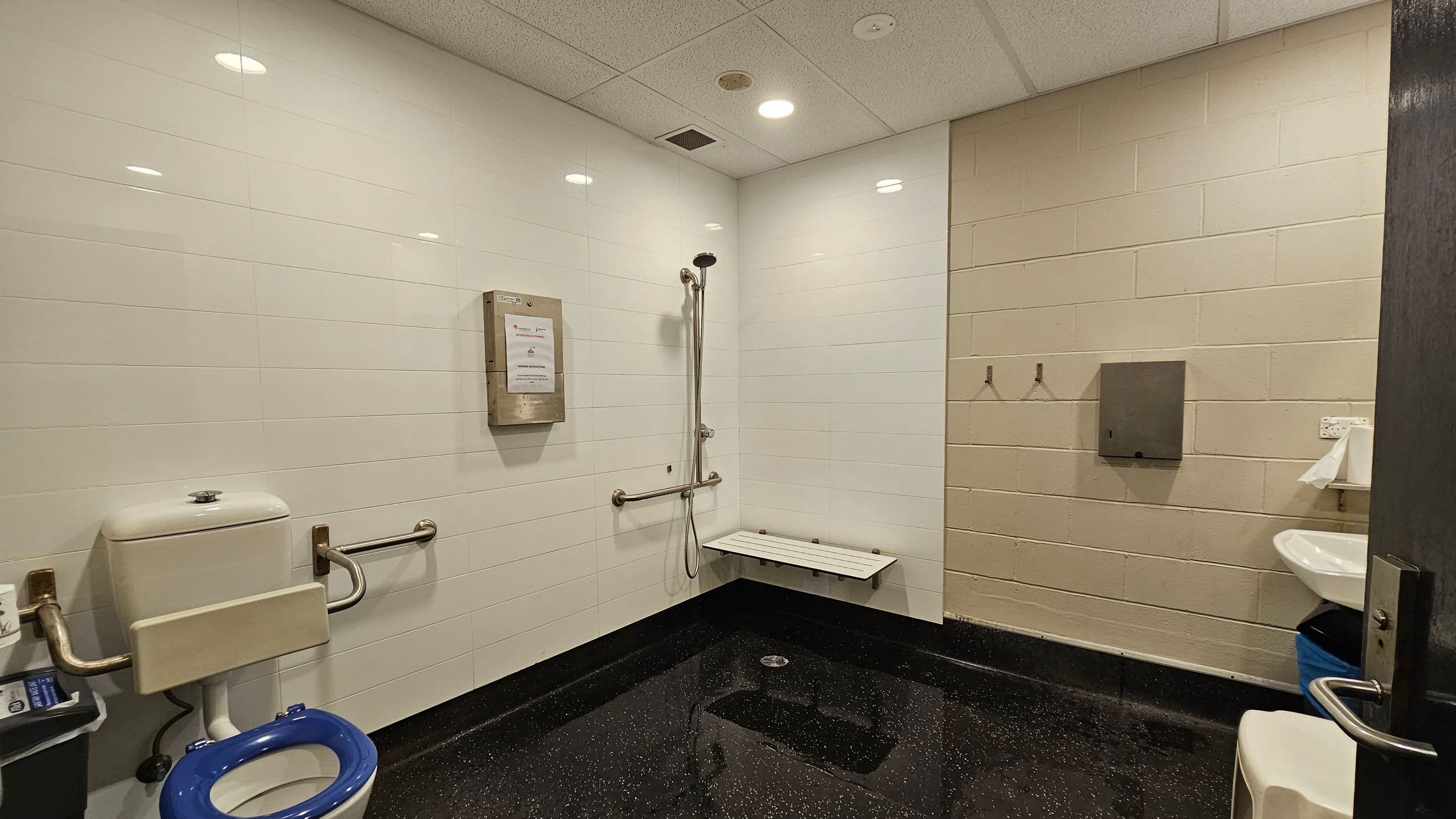 A bathroom with white tiled walls and a speckled black floor. On the left, there's a toilet with a blue seat and support rails. In the centre, a wall-mounted shower with a foldable seat and grab bars. On the right, a sink with a paper towel dispenser above it. Two hooks are mounted on the wall, and a metal box is attached to the wall near the shower. The ceiling is fitted with recessed lights and a vent.