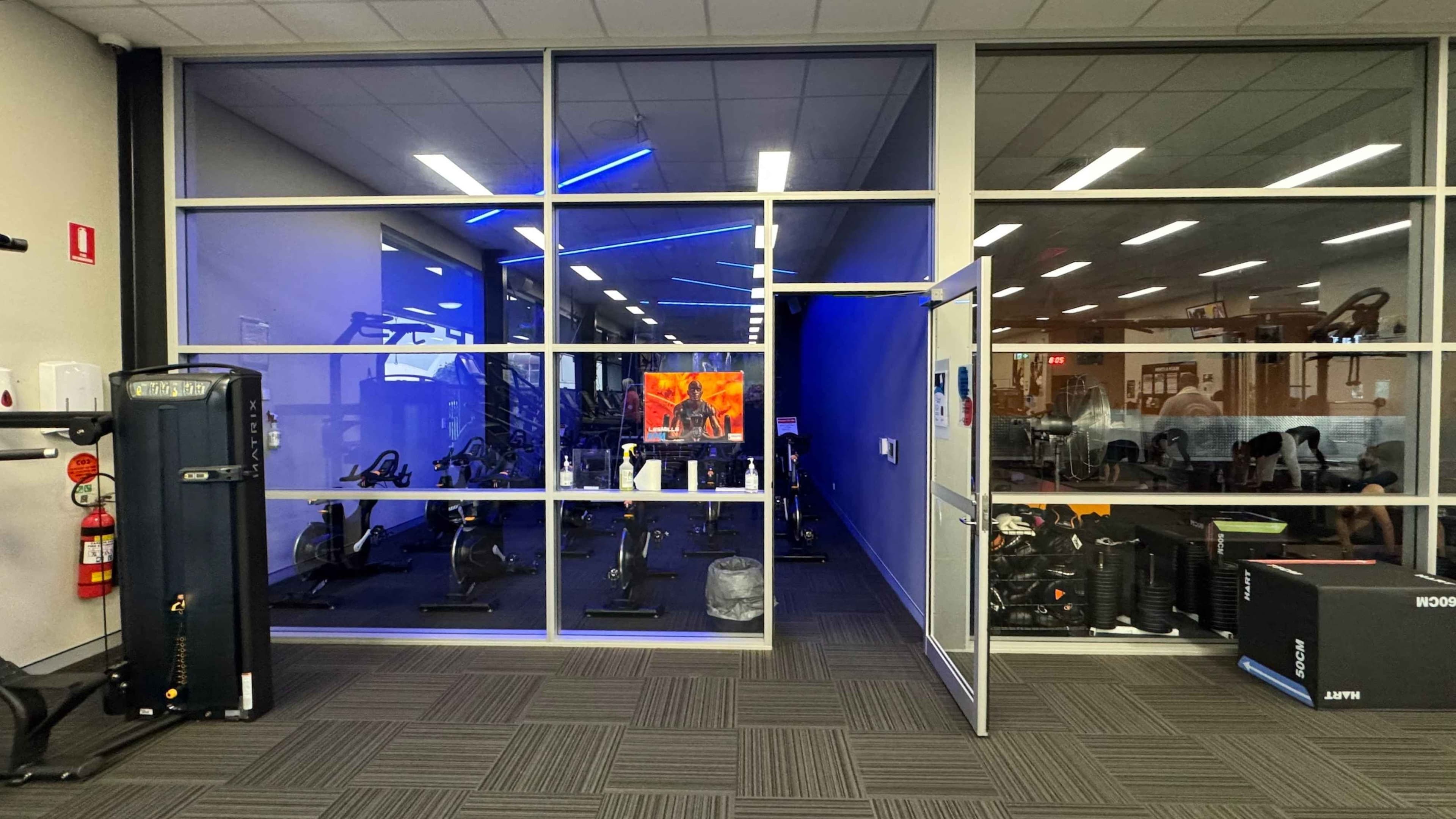A gym room with a glass wall and door. On the left, there's a black exercise machine and a fire extinguisher. In the middle, through the glass, several stationary bikes are visible, with a TV screen displaying an image of a person. The room has blue lighting. On the right, the door is open, and there are more gym equipment and a large fan. The floor is covered with green patterned carpet tiles, and the ceiling has white panels with fluorescent lights.