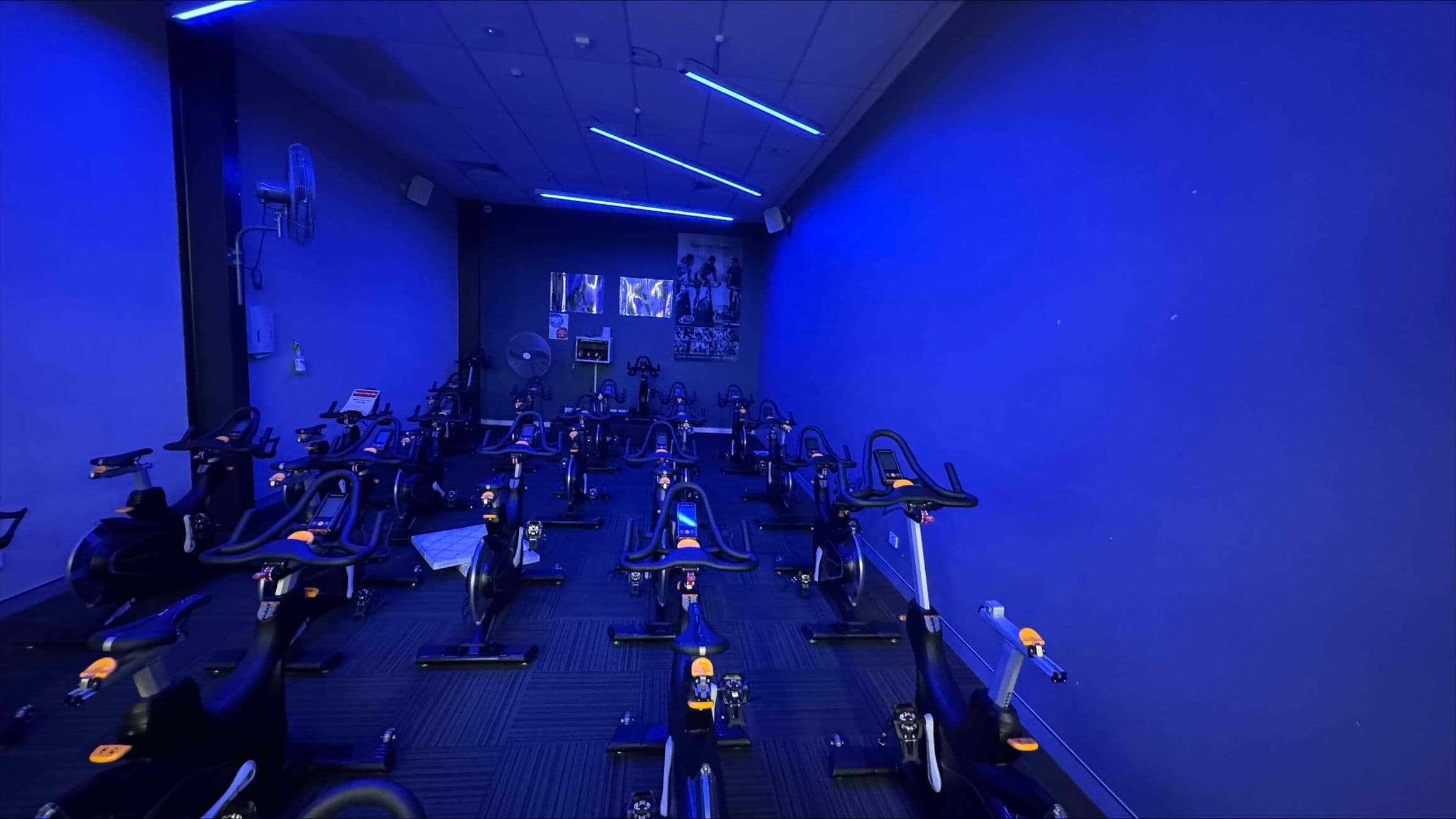 A dimly lit indoor cycling studio with a blue hue. On the left, several stationary bikes are lined up, with a wall-mounted fan above them. The right side also features more bikes, all facing the front of the room. The back wall has posters and a control panel. The ceiling has blue LED strip lights, and the floor is carpeted with a dark pattern.