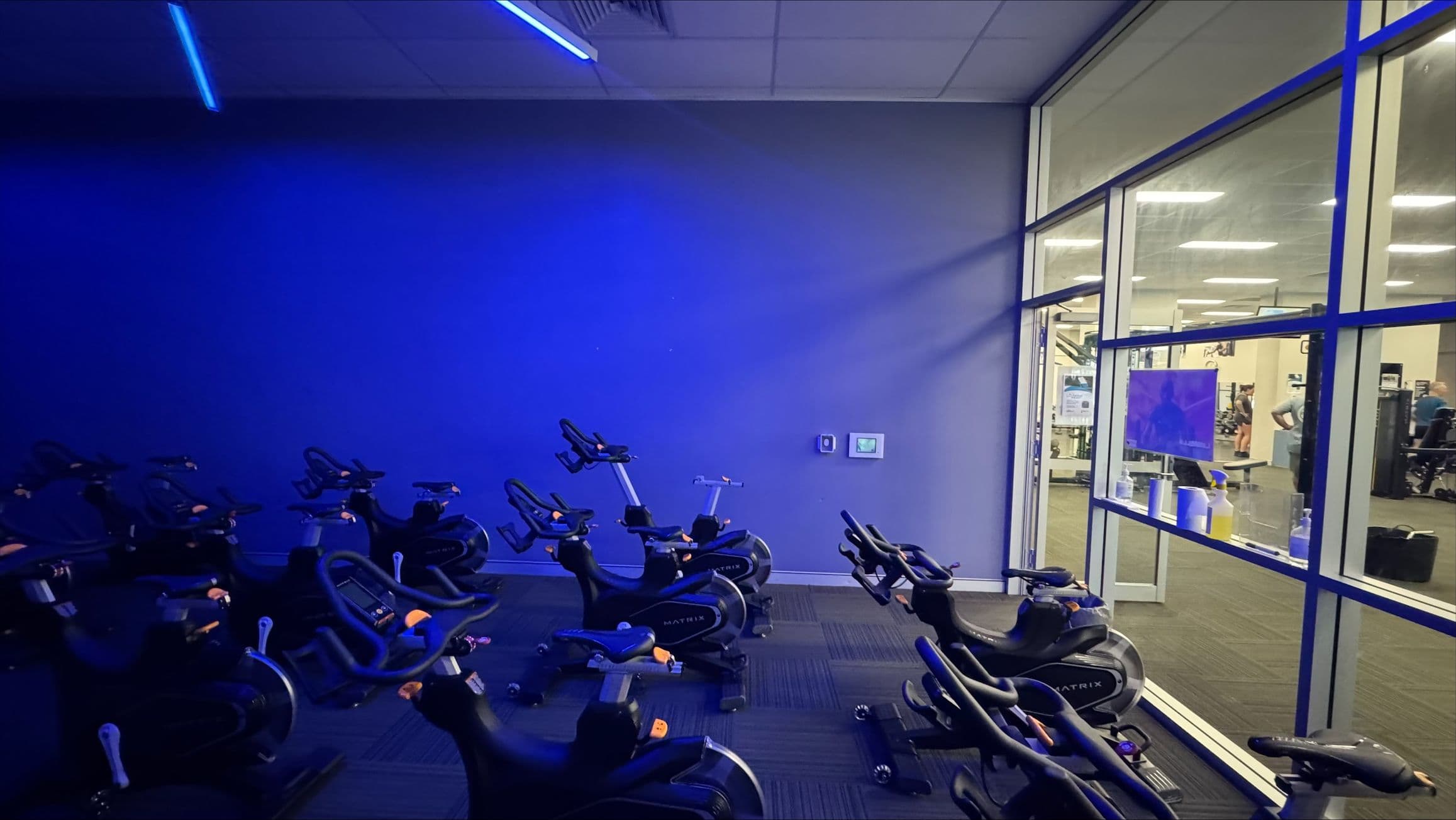 A dimly lit indoor cycling room with several stationary bikes arranged in rows on a dark carpeted floor. The left side of the room is illuminated with blue lighting, casting a glow on the bikes. On the right, a large glass wall separates the cycling area from a brightly lit gym space, where exercise equipment and people are visible. The ceiling has recessed lighting and a blue light strip.