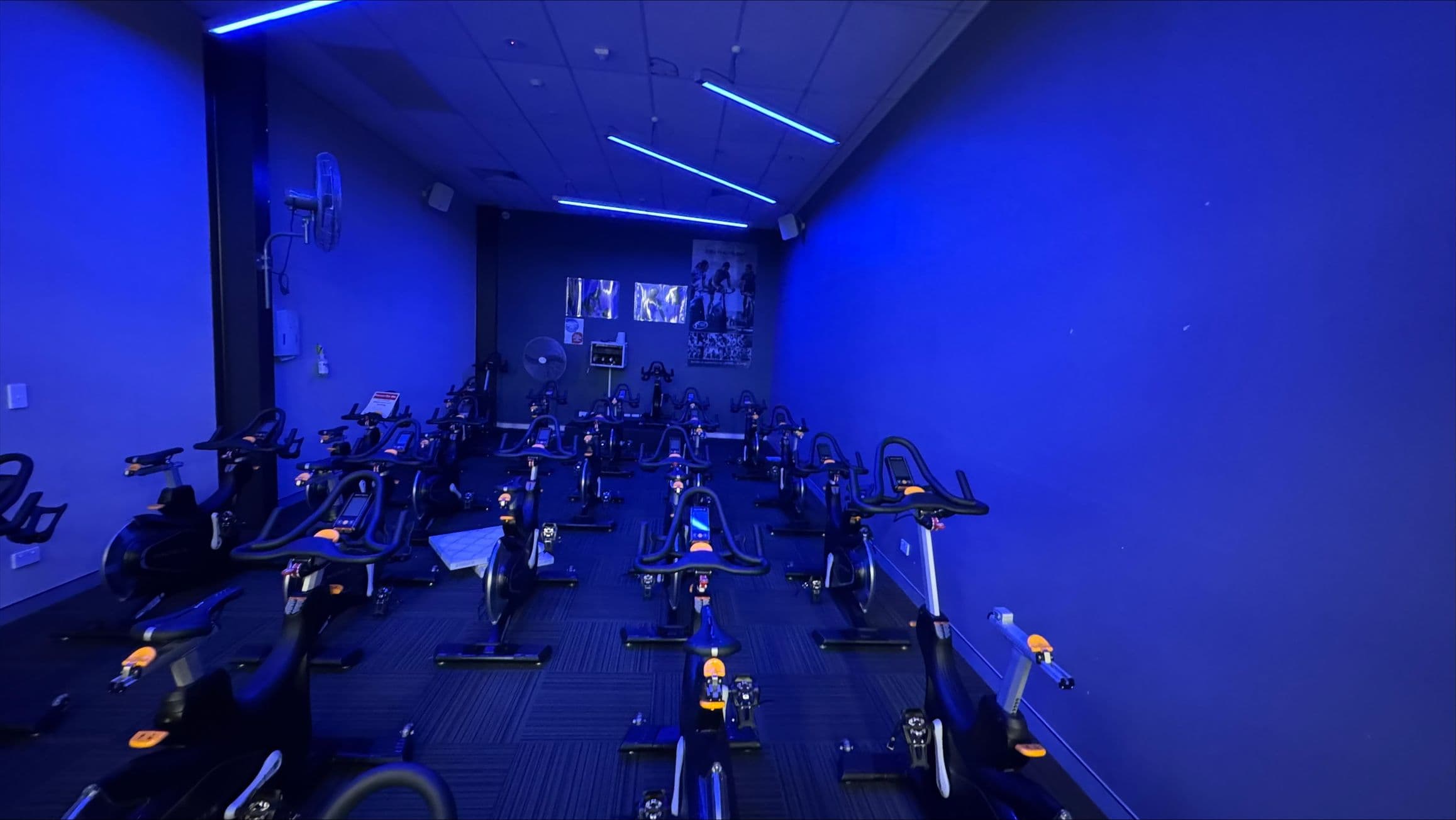 A dimly lit indoor cycling studio with a blue hue. The room is filled with stationary bikes arranged in rows. The walls are painted blue, and there are posters on the far wall. The ceiling has blue LED strip lights. A fan is mounted on the left wall. The floor is carpeted with a dark pattern.