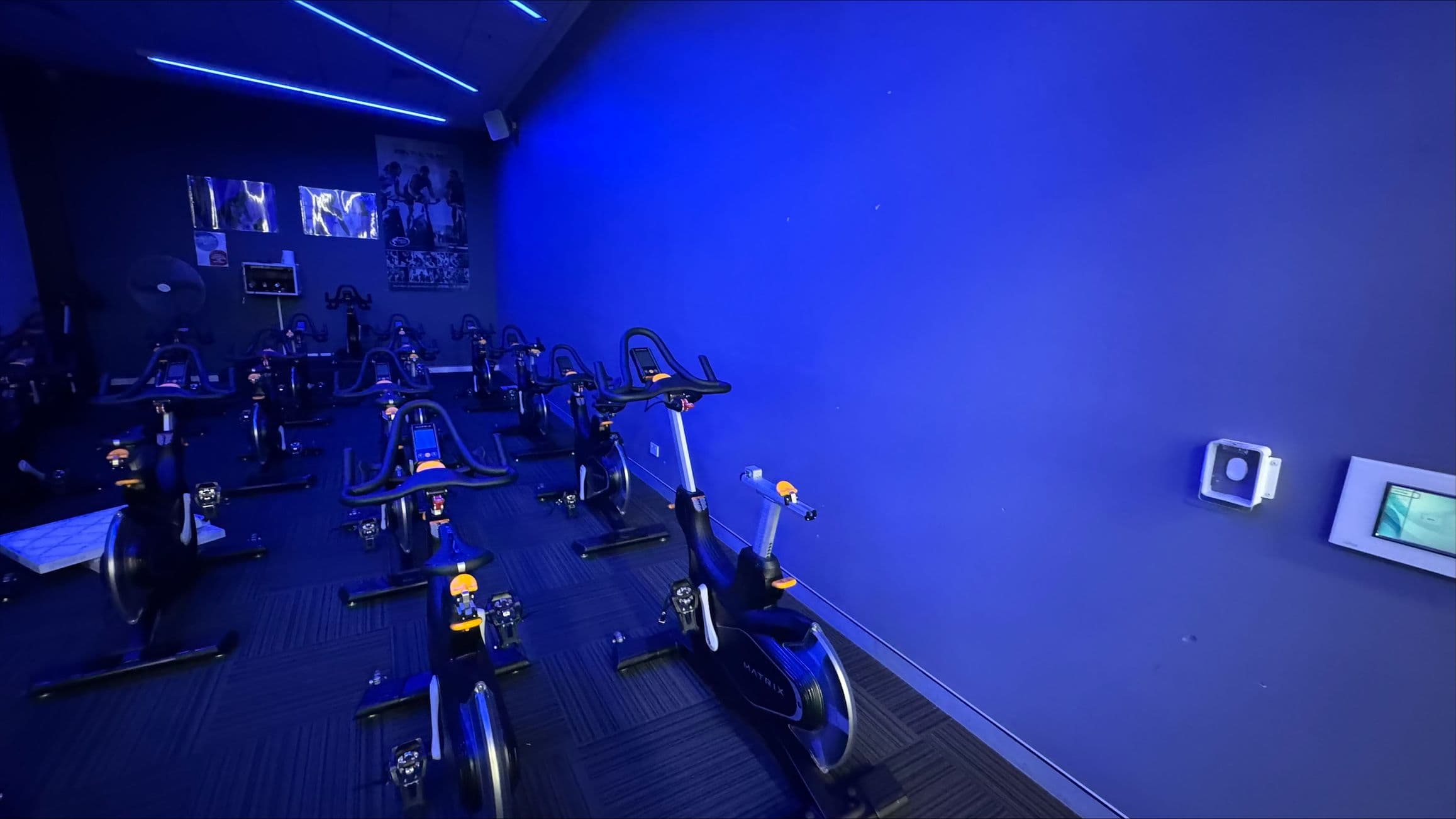 A dimly lit indoor cycling studio with a blue hue. On the left, several stationary bikes are arranged in rows, facing a dark wall with posters and a screen. The right side features a plain blue wall with a small control panel and a sensor. The ceiling has blue LED strip lights, and the floor is covered with dark carpet tiles.
