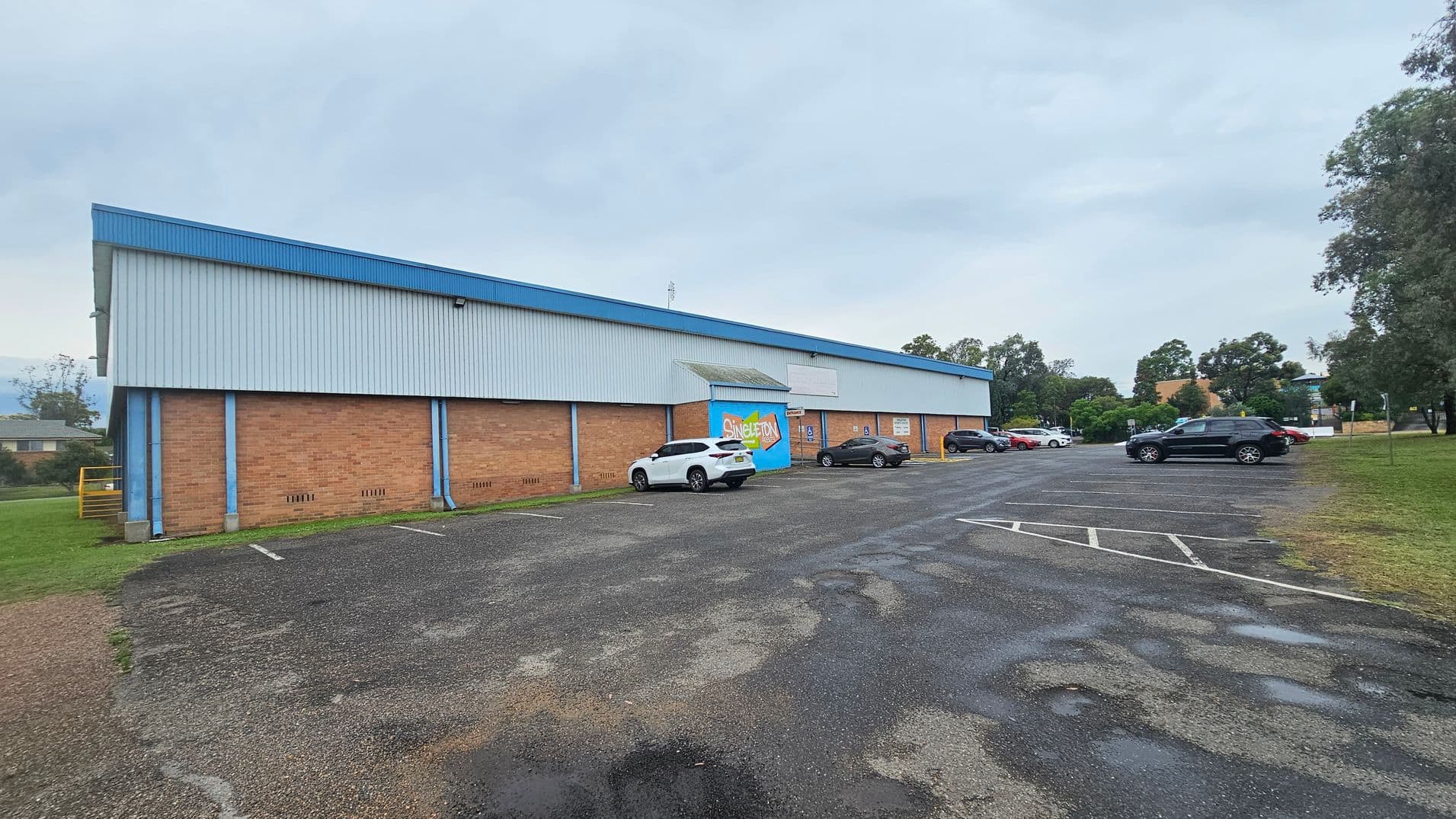A large rectangular building with a brick lower half and a corrugated metal upper half, painted in blue and grey. The front has a mural with the word "Singleton" and several parked cars, including a white SUV and a black car. The car park is mostly empty, with marked parking spaces on an asphalt surface. Grass and trees are visible on the right side, and a small patch of grass is on the left.
