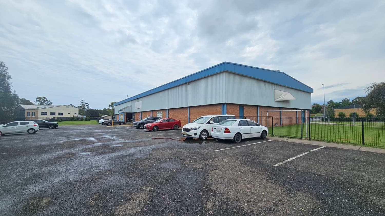A large, rectangular building with a blue roof and brick walls is on the right. Several cars are parked in front of it on an asphalt surface. To the left, there is a smaller building in the background, surrounded by grass and trees. A metal fence runs along the right side, enclosing a grassy area. The sky is overcast.