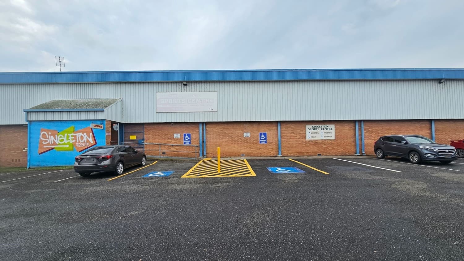 A large building with a blue and brick exterior. On the left, there's a mural with "Singleton Heights" painted in white and orange on a blue background. Two cars are parked in front, one on the left and one on the right, with designated disabled parking spaces marked in blue and yellow. A sign on the right reads "Singleton Sports Centre" with sports listed below. The ground is asphalt, and the building has a corrugated metal roof.