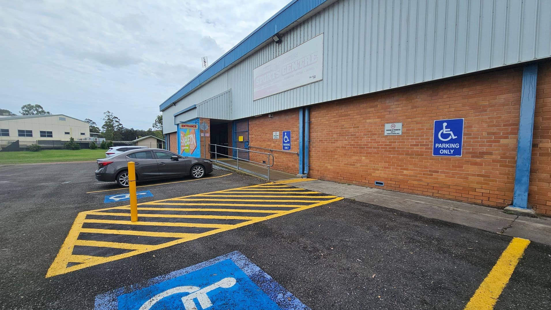 A parking area with a designated disabled parking space marked in blue on the ground. A grey car is parked nearby. The building on the right has a brick wall with a blue and white "Parking Only" sign. A ramp with railings leads to an entrance with a colourful sign. The building has a corrugated metal roof. In the background, there is another building and some trees. The ground is asphalt, and there are yellow lines marking the parking area.