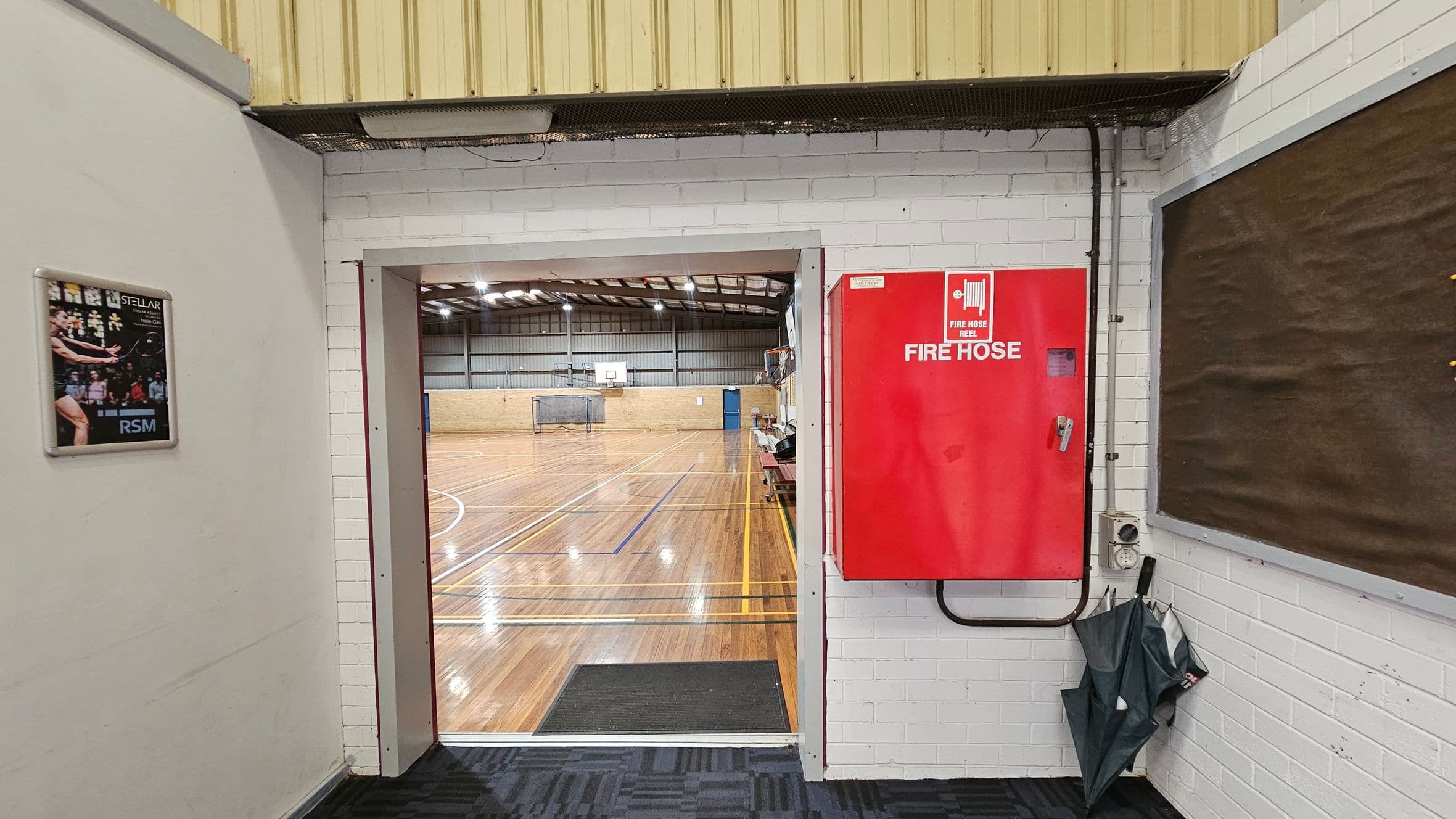 A doorway leads to an indoor court with a wooden floor and basketball hoops. On the left wall, there's a framed poster. On the right, a red fire hose cabinet is mounted on the white brick wall, with umbrellas stored below. The ceiling is metal with exposed beams. The floor in the foreground is carpeted.