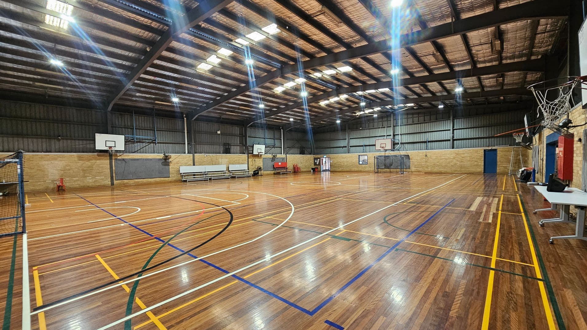 A spacious indoor sports hall with a polished wooden floor marked for various sports. On the left, there are basketball hoops and a net. In the centre, several benches are lined up against the wall. On the right, more basketball hoops and a few tables are visible. The ceiling is high with exposed beams and bright overhead lights. The walls are made of brick and metal panelling, with a few doors and sports equipment scattered around.