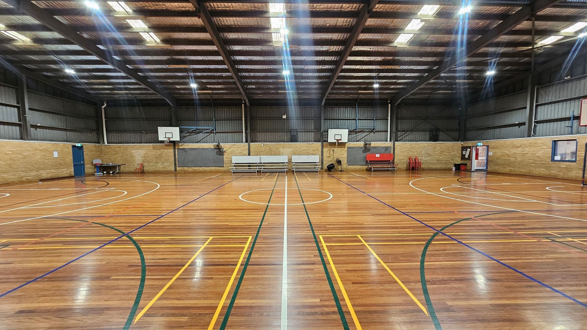 A spacious indoor basketball court with a polished wooden floor marked with various coloured lines. Two basketball hoops are mounted on the far wall. Benches and a table are positioned against the wall, with a stack of red chairs nearby. The walls are made of brick and metal panelling. The ceiling is high with exposed beams and fluorescent lights. A blue door is on the left, and a window and another door are on the right.