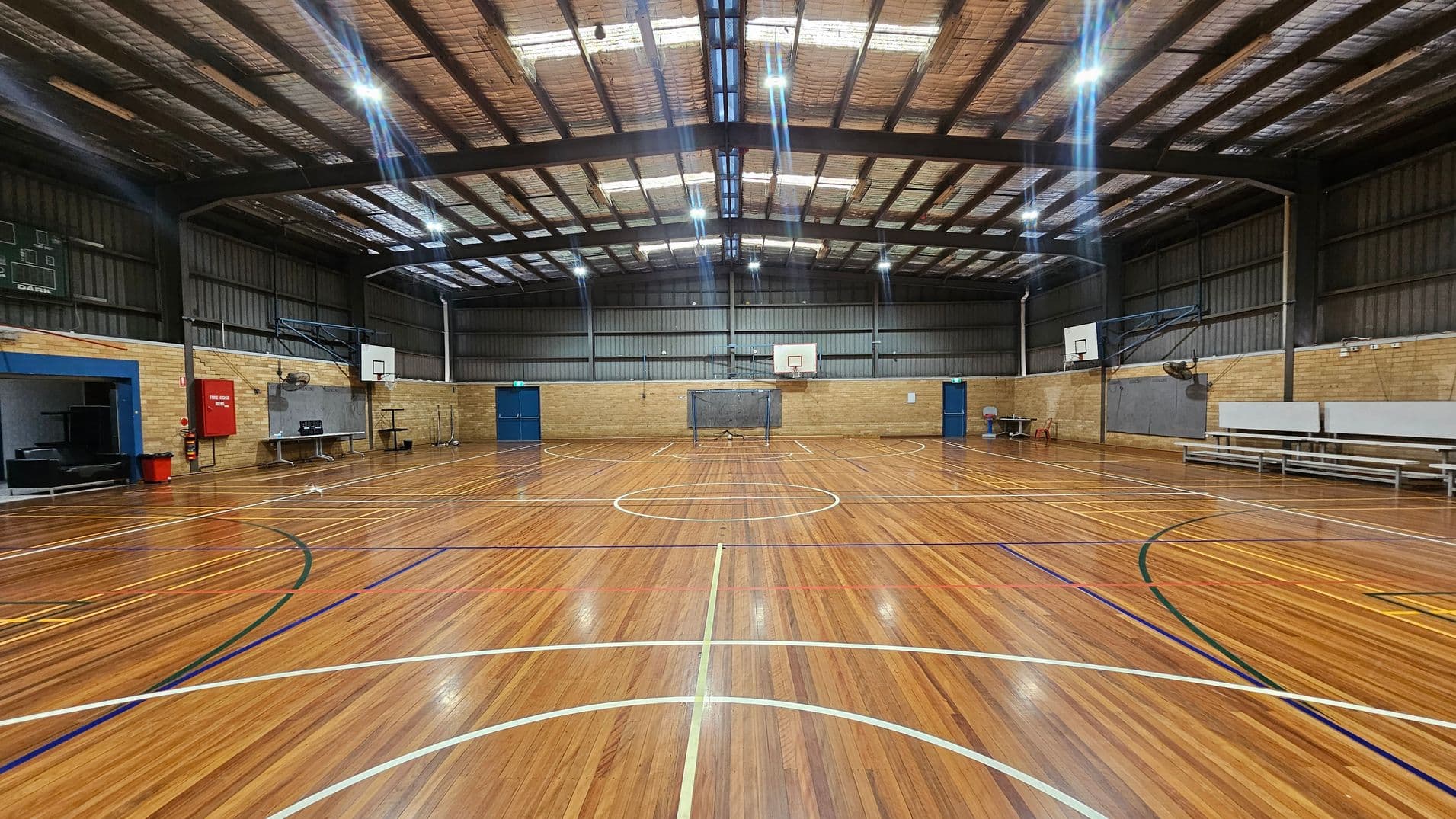 A spacious indoor basketball court with a polished wooden floor marked with various coloured lines. Two basketball hoops are mounted on the brick walls at either end. The ceiling is high with exposed beams and bright overhead lights. On the left, there is a scoreboard and a red fire hose reel. On the right, there are benches and a fan mounted on the wall. Blue doors are visible on both sides of the court.