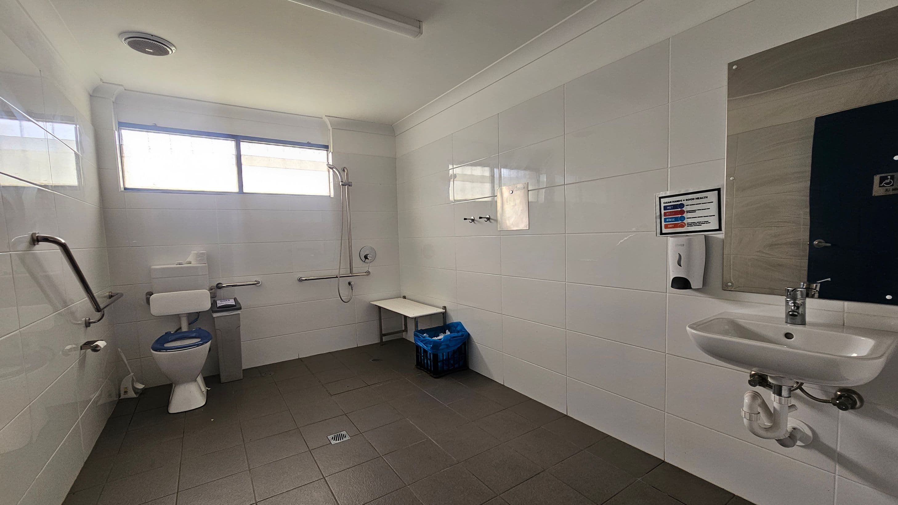 A spacious, accessible bathroom with white tiled walls and a grey tiled floor. On the left, there is a toilet with support rails and a toilet paper holder. Above the toilet, a window allows natural light in. In the centre, a shower area with a wall-mounted showerhead and a foldable bench. On the right, a sink with a mirror above it, a soap dispenser, and a sign with instructions. A blue bin is placed under the bench. The ceiling is white with a vent.