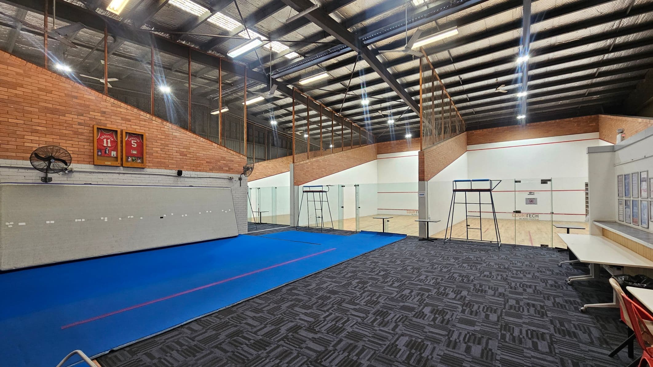 A sports facility with a blue mat on the left and a squash court on the right. The floor is covered with patterned carpet tiles. The ceiling is high with exposed beams and fluorescent lights. On the left wall, there are framed sports jerseys and a wall fan. A metal ladder stands near the glass-walled squash court. Tables and chairs are positioned on the right side. The walls are a mix of brick and white surfaces.