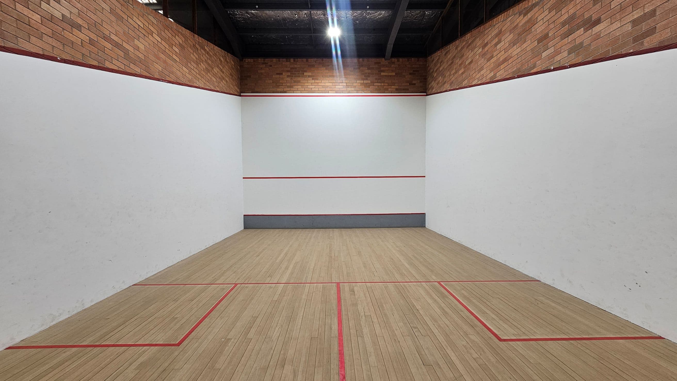 A squash court with white walls and a wooden floor marked with red lines. The upper section of the walls is made of exposed brick. The ceiling is dark with visible beams and lighting fixtures. The front wall has a red line across it, and the floor is divided into sections by red boundary lines.