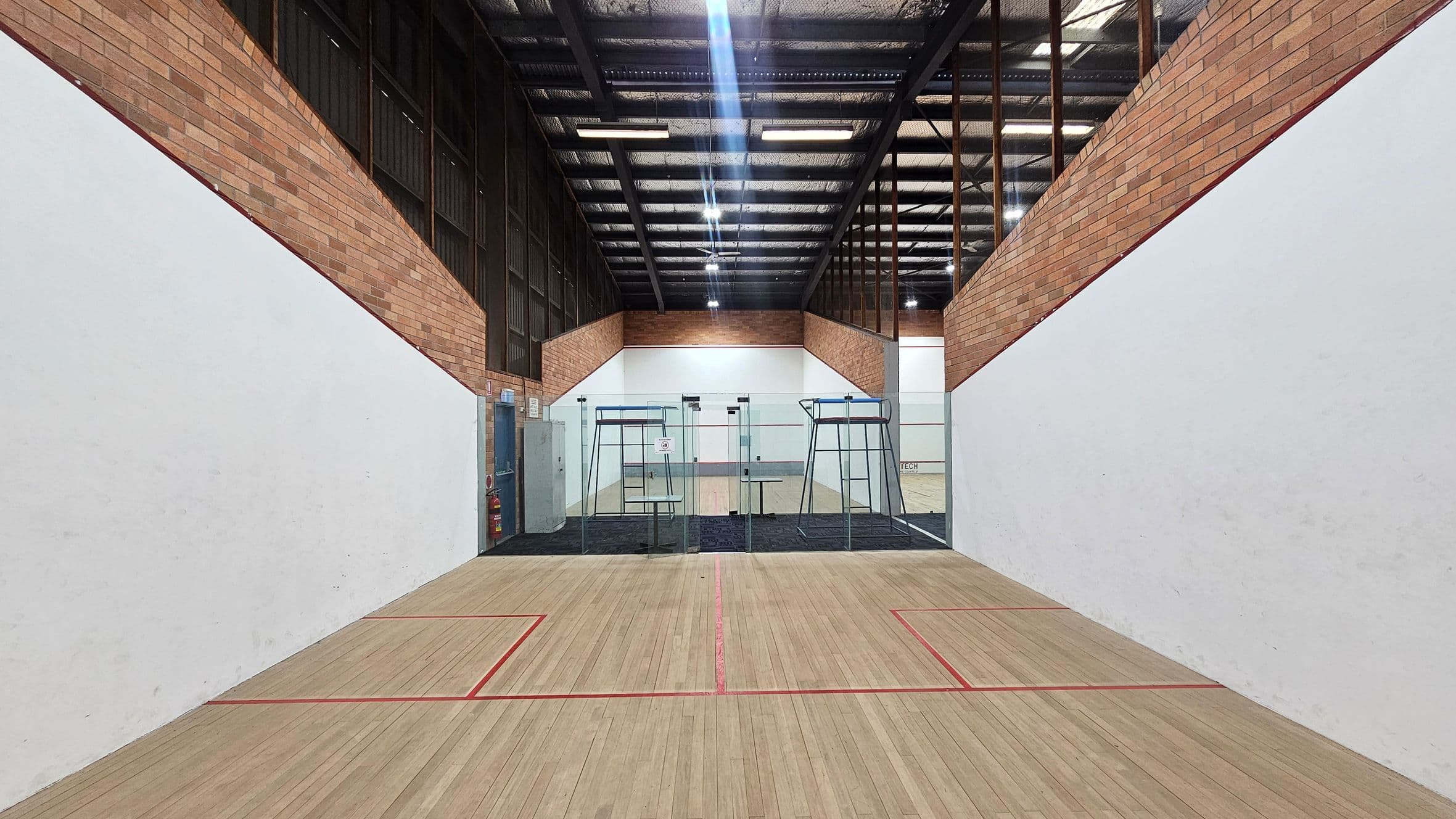 A squash court with wooden flooring marked by red lines. The walls are white with exposed brick at the top. At the far end, there is a glass wall separating the court from another area. Above, the ceiling is industrial with visible beams and lighting. On the left, there is a door and a fire extinguisher.
