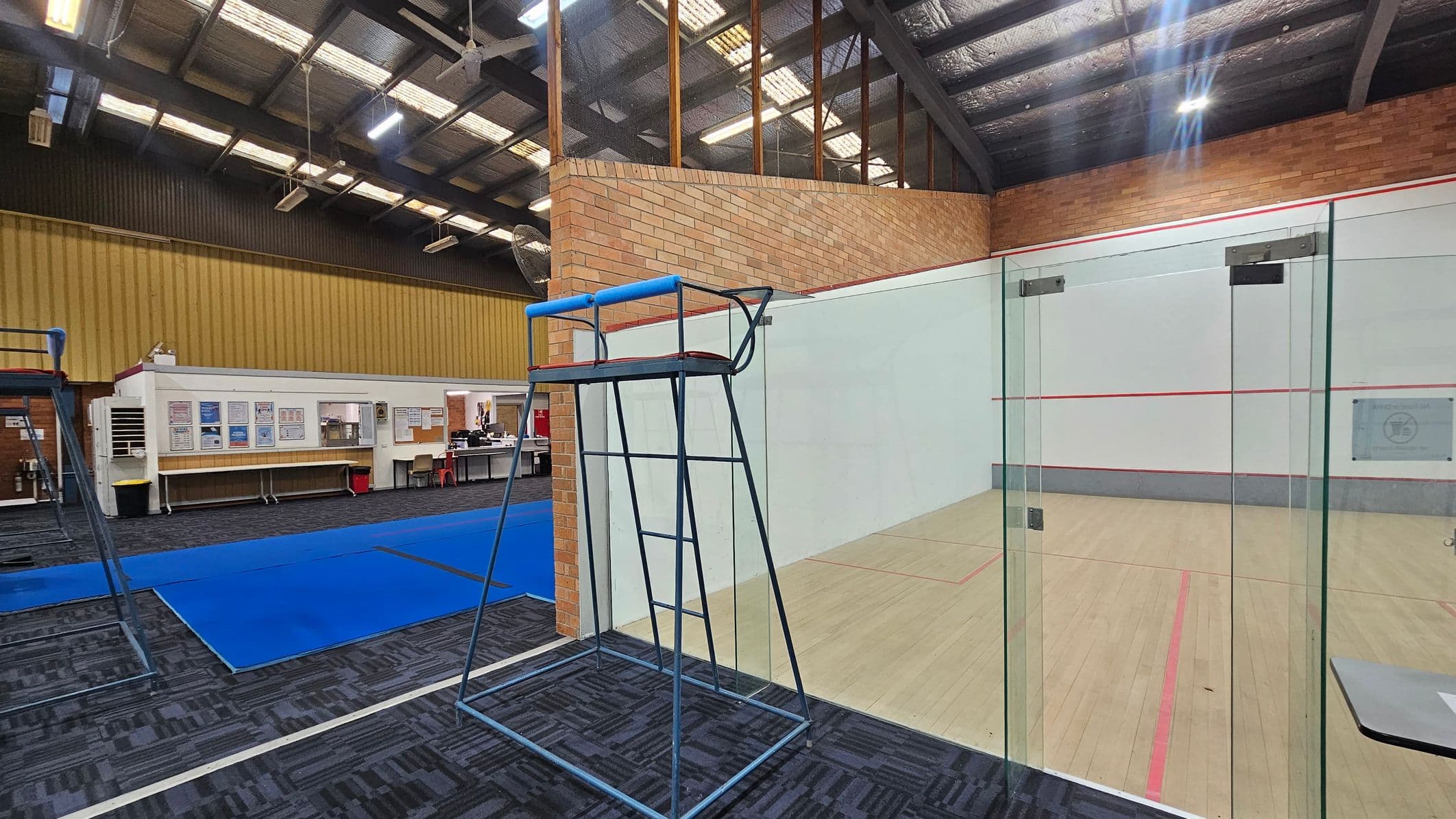 A sports facility with a squash court on the right, featuring glass doors and a wooden floor marked with red lines. In the foreground, a blue referee chair stands on a patterned carpet. On the left, a blue mat covers part of the floor, and a white office area with desks and chairs is visible. The ceiling is high with exposed beams and lights, and the walls are made of brick and corrugated metal.