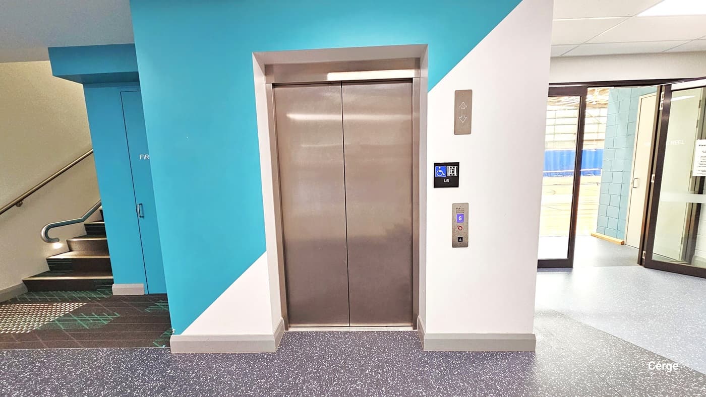 Access Level One through the stairs on the left or the lift. The lift has up and down buttons for operation. A blue door by the stairs is the Fire Exit, while a partially open glass door on the right leads to a court. Well-lit space with a grey granite floor and blue and white walls by the lift.
