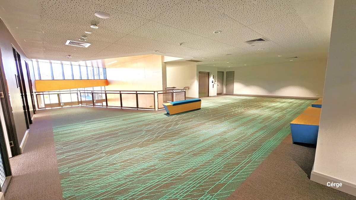 A close-up of Level One reveals a spacious area with yellow and blue wooden benches. The floor features granite with green criss-cross markings. The white ceiling has fluorescent bulbs and central air conditioning. To the right, three doors lead to the lift, stairs, and fire exit. To the left, is an open space with floor-to-ceiling glass windows.
