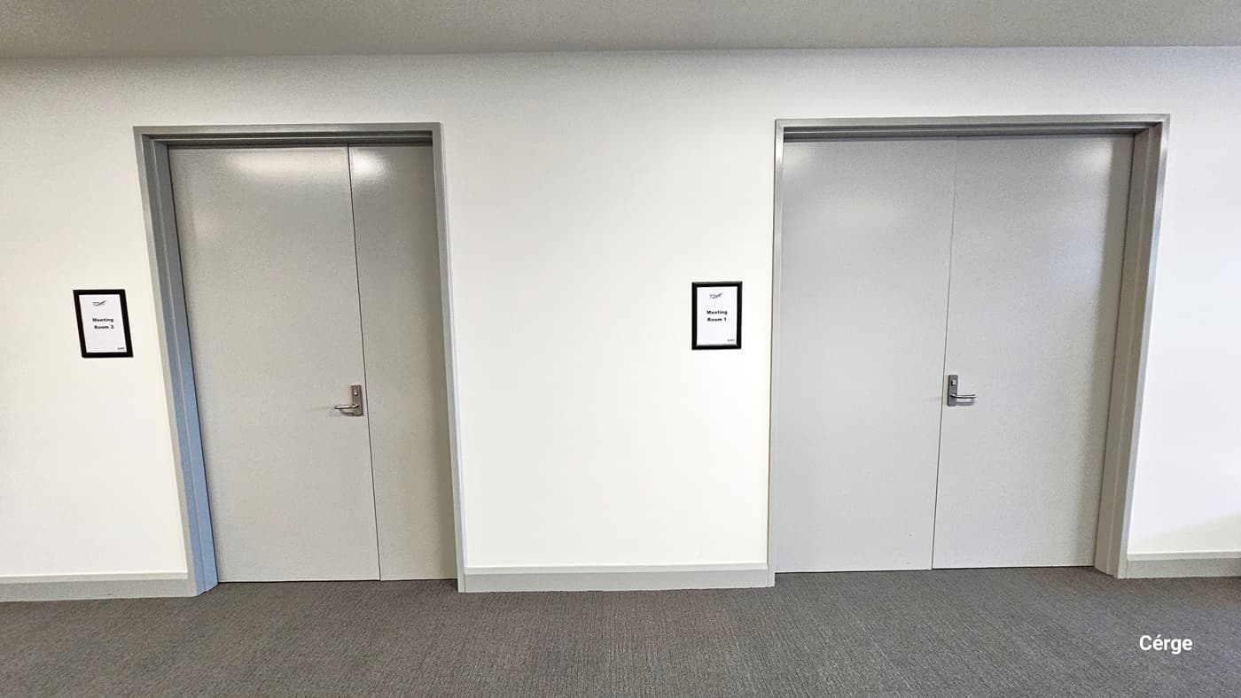 Entry to the meeting rooms. On Level One, this area features  two grey-painted doors, each with stainless steel handles. These doors lead to Meeting Rooms, with labels enclosed in black and white frames. The walls are plain white, and the floor has a grey carpet-like design.