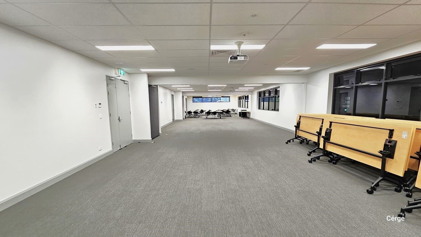 The lobby area offers a spacious setting with white walls and ceilings. Fluorescent lights, and a central air conditioning are ceiling-mounted. On the right, yellow foldable and mobile tables align with long brown glass windows. To the left, a grey fire exit door and  a couple of grey doors. Towards the lobby's end, neatly arranged tables, chairs, and four flat screens. The floor has a grey, carpet-like finish.