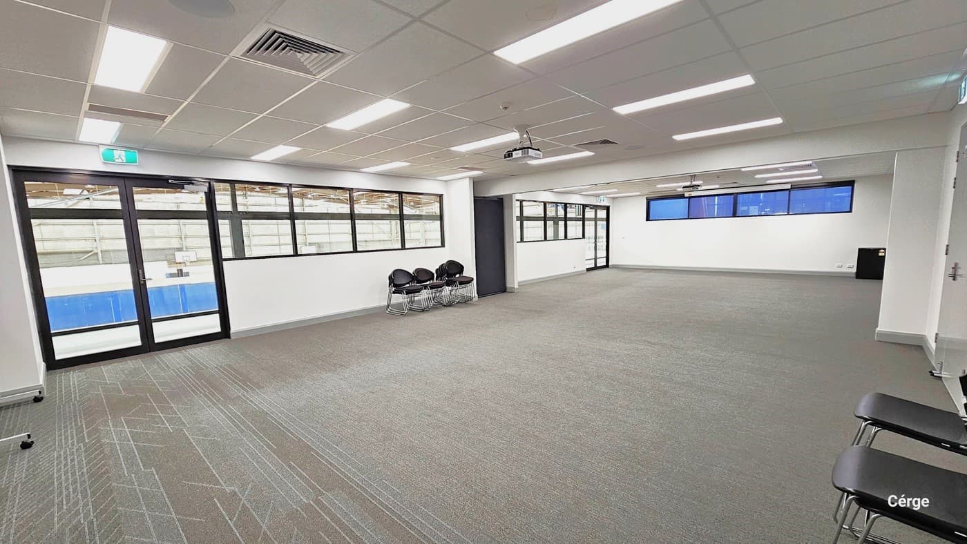 Within the meeting room, the scene presents a roomy area with white walls and ceilings. Ceiling-mounted features include fluorescent lights, and central air conditioning. On the right, two chairs are arranged. To the left, there's a glass door, brown-framed glass windows, and a row of black chairs against the wall. The floor exhibits a grey, crisscross design with a carpet-like finish.