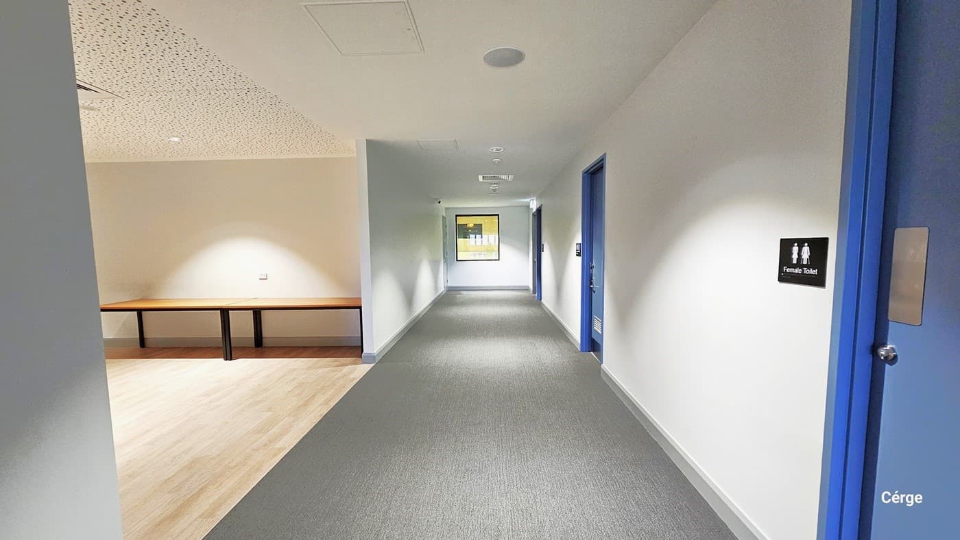 This is a vew of the corridor outside a meeting room, with grey carpeted flooring. To the left is an open space with timber flooring and to the right is access to the unisex toilet.