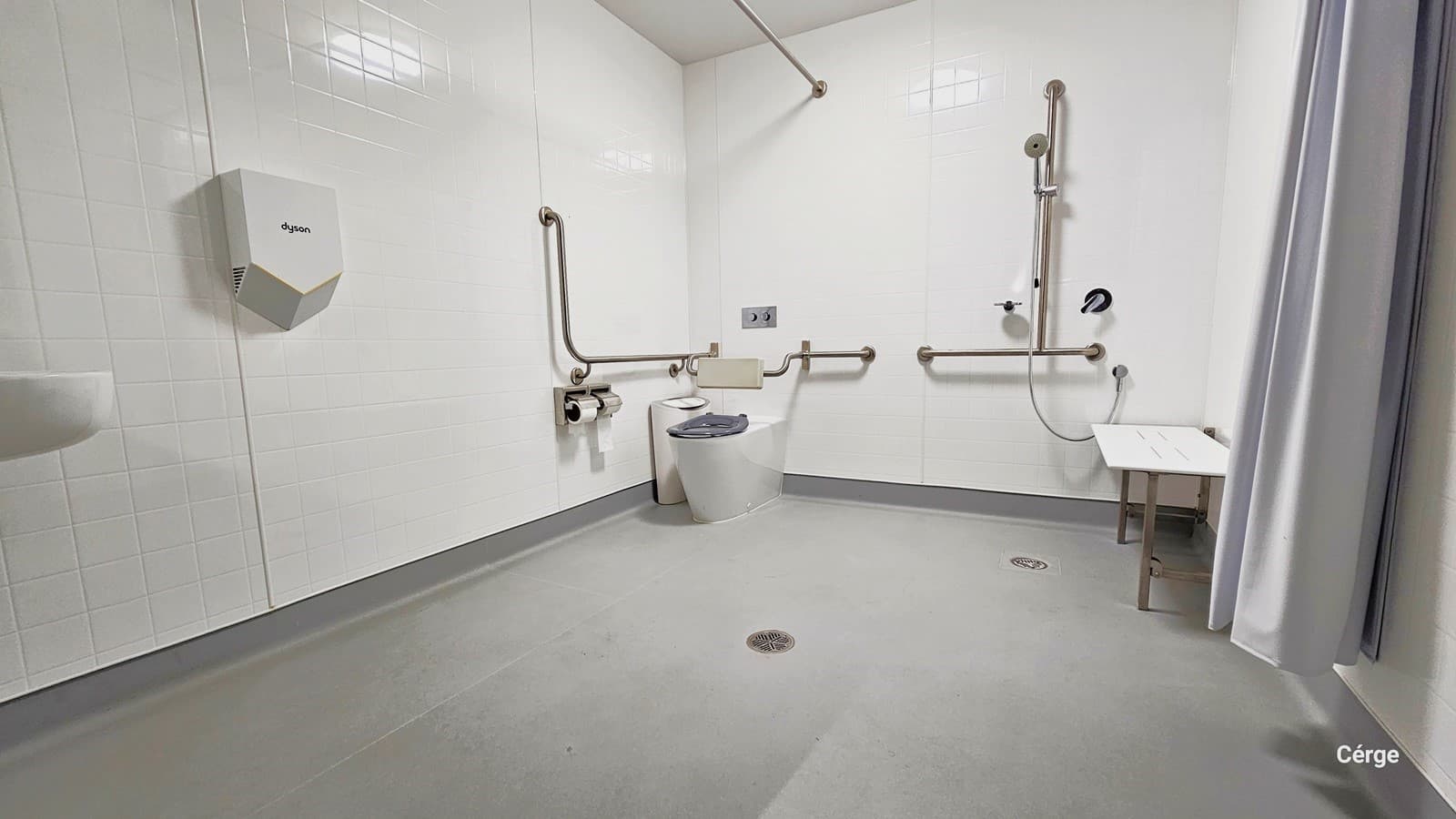 Within the bathroom, the space is well-lit, featuring white brick-patterned walls and a white ceiling. On the left, is a washbasin and hand dryer. In the corner, you see a white ambulatory toilet, rubbish bin, and tissue dispenser. Mobility aids include a couple of handrails, a white bench, a shower, and a curtain. The floor has a matte grey finish.