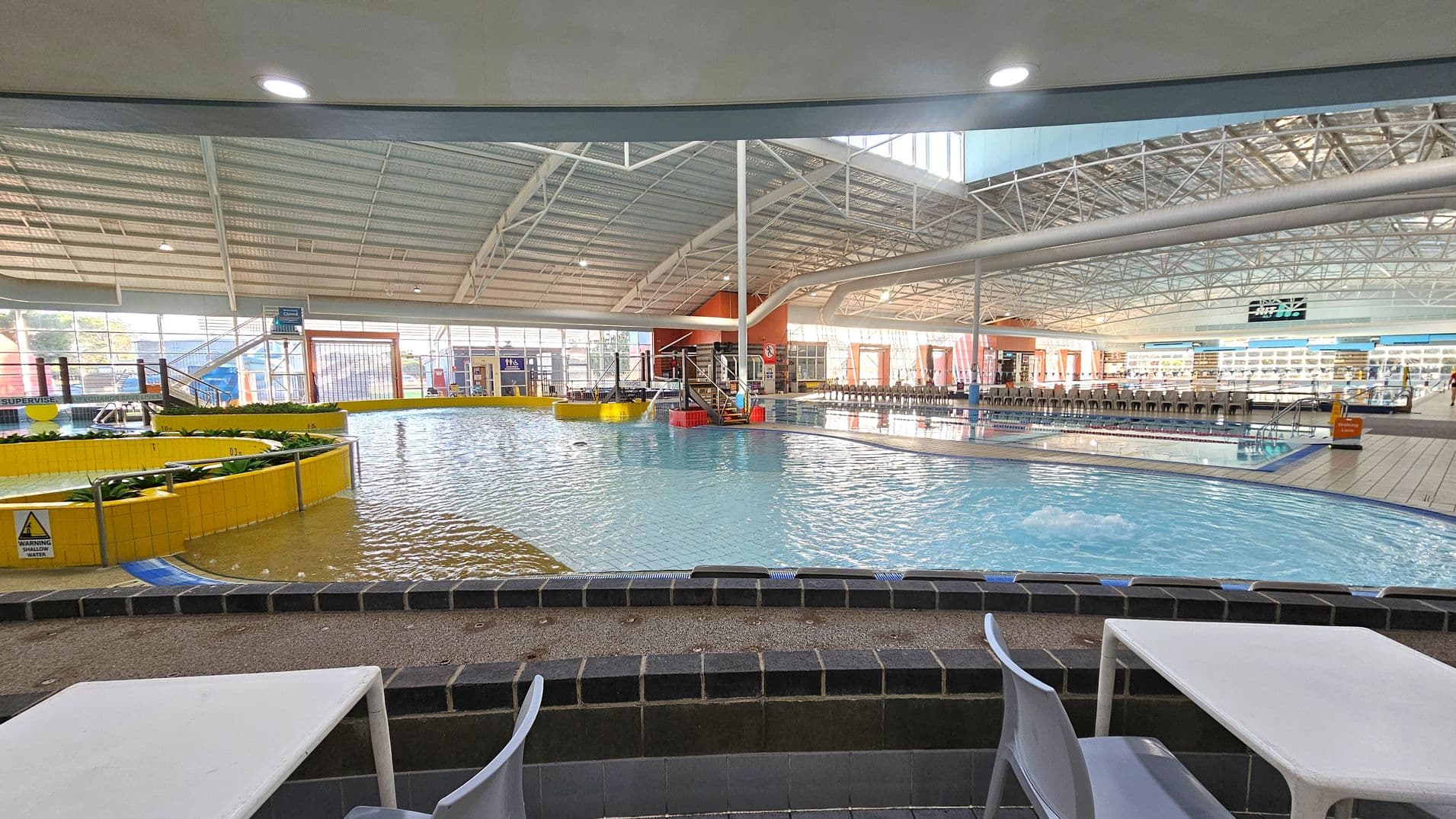 Indoor swimming pool area with a shallow pool in the foreground, surrounded by a low black-tiled wall. White tables and chairs are positioned near the pool. The pool has a yellow-tiled circular feature with plants. In the background, there are more pools, a slide, and a row of chairs. The ceiling is high with metal beams and skylights, allowing natural light to illuminate the space.
