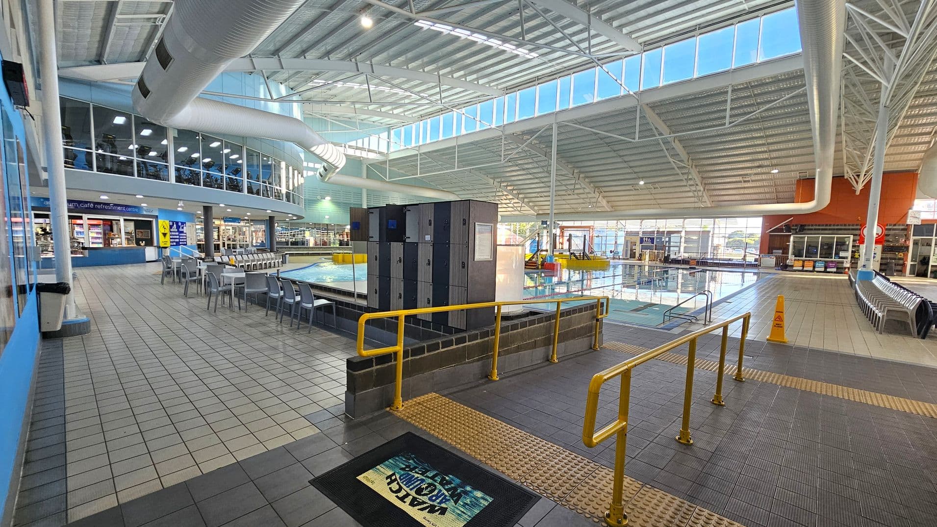 An indoor aquatic centre with a large pool on the right, surrounded by tiled flooring. In the foreground, there are yellow railings and a mat with a warning sign. Lockers are positioned near the pool. On the left, a café area with tables and chairs is visible, and above it, a mezzanine with exercise equipment. The ceiling is high with exposed beams and large windows allowing natural light to enter.