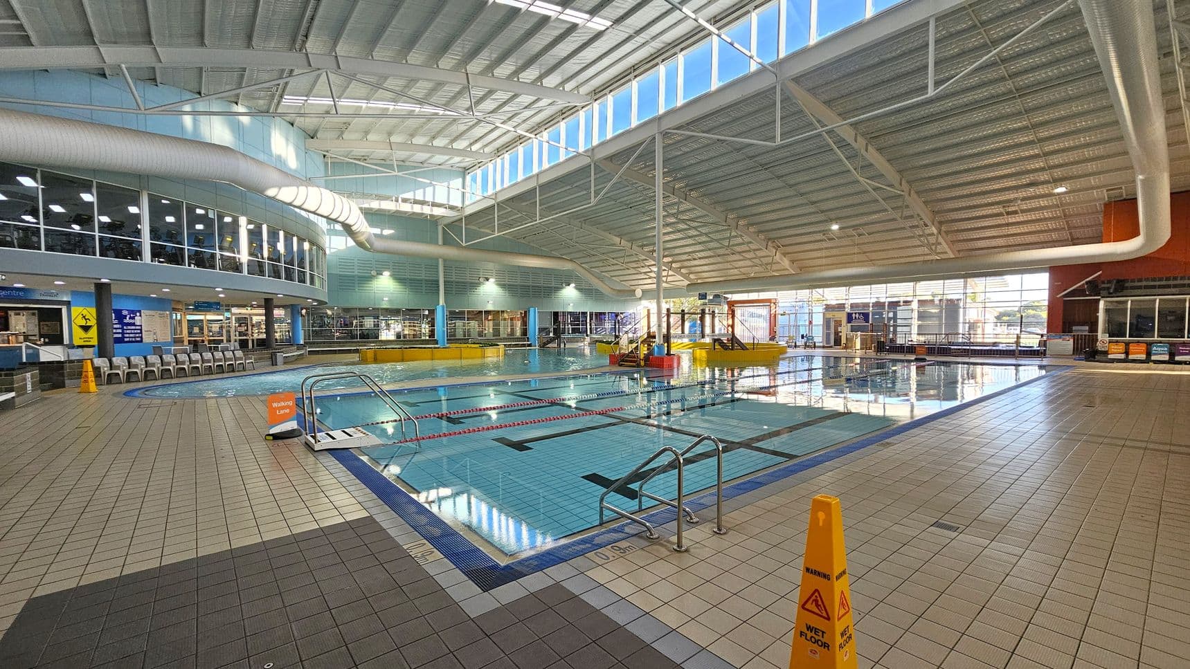 An indoor swimming pool area with a tiled floor and a high, curved ceiling featuring large windows allowing natural light. On the left, there are rows of chairs and a glass-walled gym area above. In the centre, the pool has marked lanes and a small set of steps leading into the water. On the right, there are more windows and a play area with water features. Yellow caution signs are placed around the pool, indicating a wet floor.