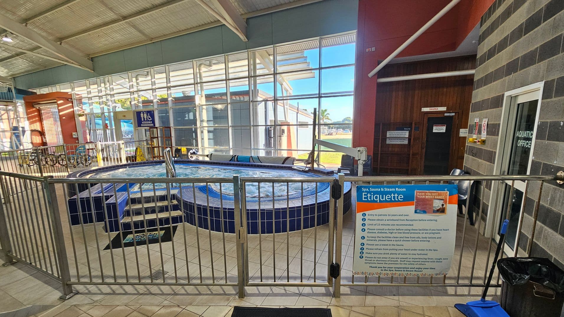 A spa area with a circular hot tub surrounded by a metal fence. On the right, a sign displays spa etiquette rules. Behind the fence, a door marked "Sauna" is visible. The left side shows large windows with a view of the outside, allowing natural light to illuminate the space. The floor is tiled, and a blue dustpan and brush are placed near the fence. The ceiling is made of metal panels with exposed beams.