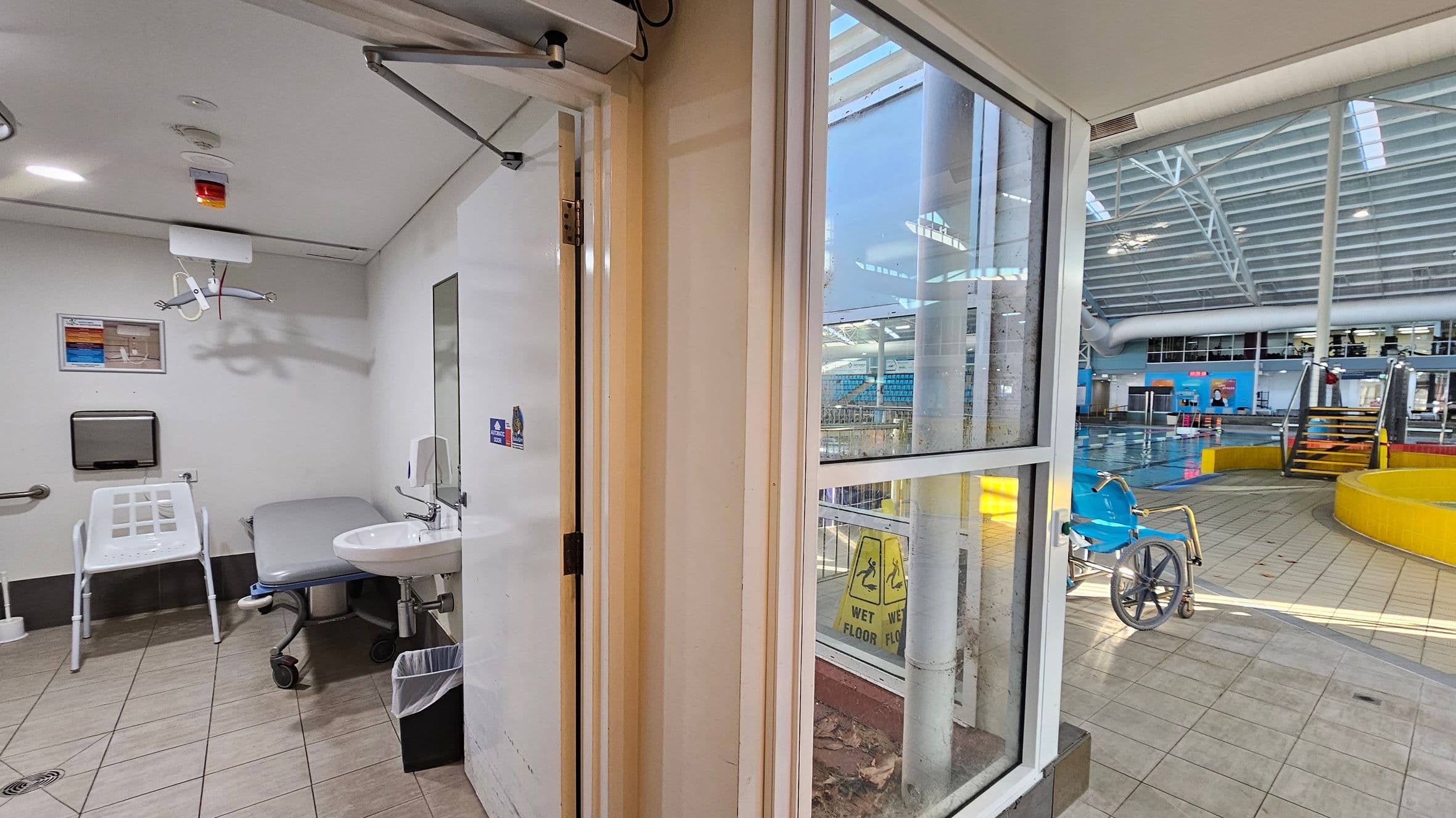 On the left, a changing room with a white chair, a sink, a mirror, and a paper towel dispenser. The ceiling has a hoist system. On the right, a swimming pool area with a blue wheelchair, yellow barriers, and steps leading into the pool. The floor is tiled, and large windows separate the two areas. A "Wet Floor" sign is visible through the window. The ceiling in the pool area has a grid-like structure with skylights.