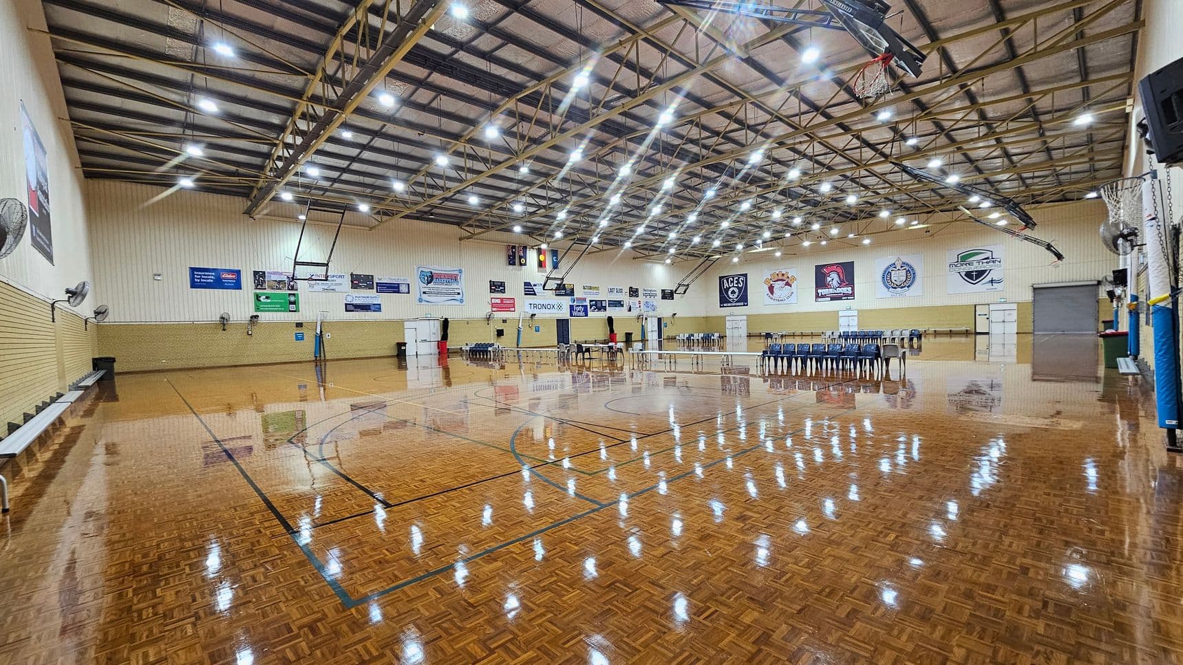 A spacious indoor sports hall with a polished wooden floor marked for basketball. On the left, there are benches and wall-mounted fans. The right side features a row of stacked chairs and a basketball hoop. The back wall displays various banners and logos. The ceiling is high with exposed beams and numerous bright lights.