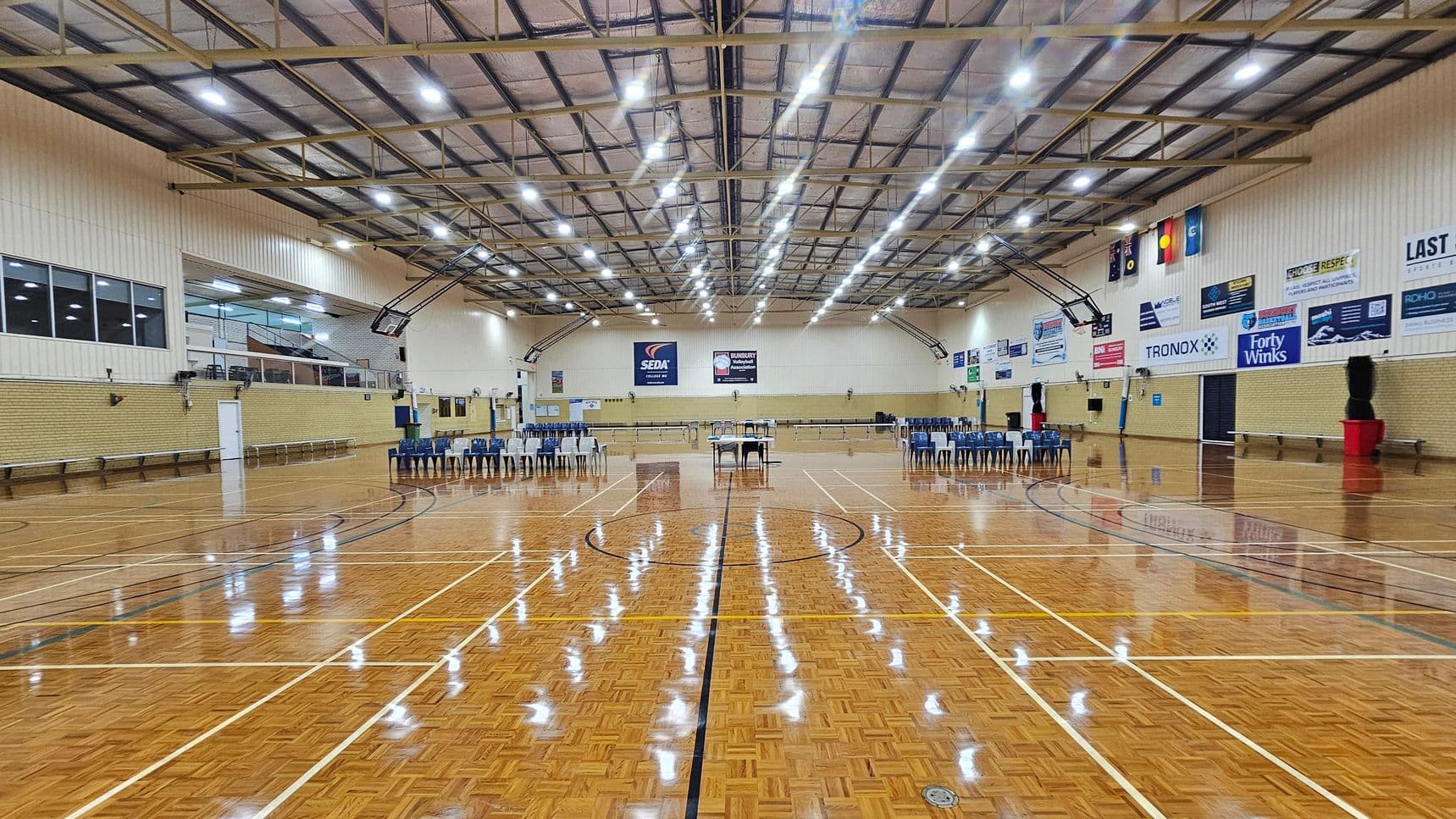 A large indoor sports hall with a polished wooden floor marked for various sports. In the centre, there are rows of blue chairs and a table. The walls are lined with banners and advertisements. The ceiling is high with exposed beams and bright overhead lights. On the left, there are windows and a small upper viewing area. On the right, more chairs are arranged, and there are flags displayed on the wall. The space is open and well-lit.