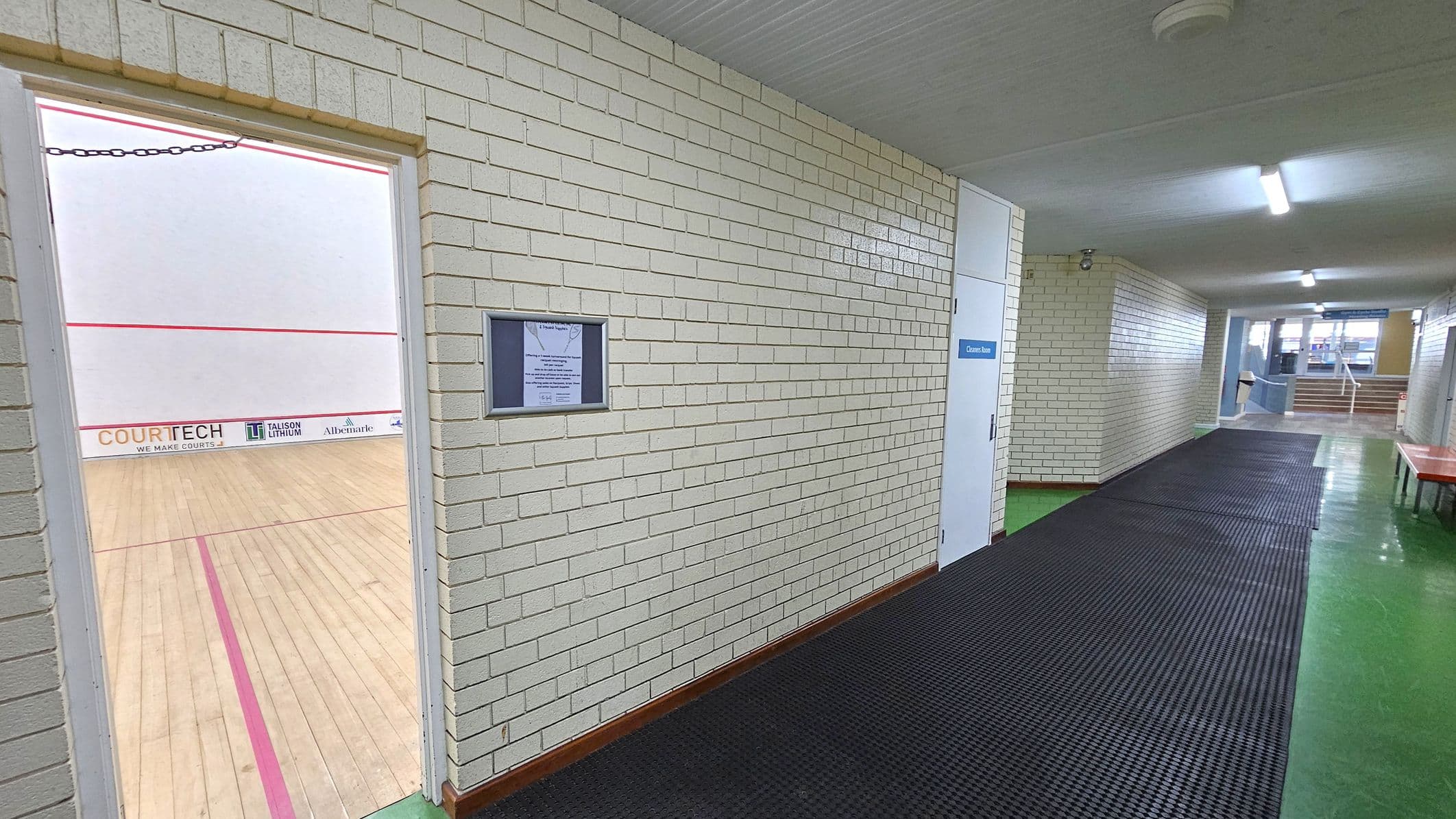 A corridor with white brick walls and a green floor. On the left, an open door reveals a squash court with a wooden floor and red boundary lines. The court has advertisements on the lower wall. On the right, a white door labelled "Cleaner Room" is visible. The corridor has a black rubber mat running along the floor. In the background, there are stairs leading up, with windows allowing natural light to enter. The ceiling is white with fluorescent lights.
