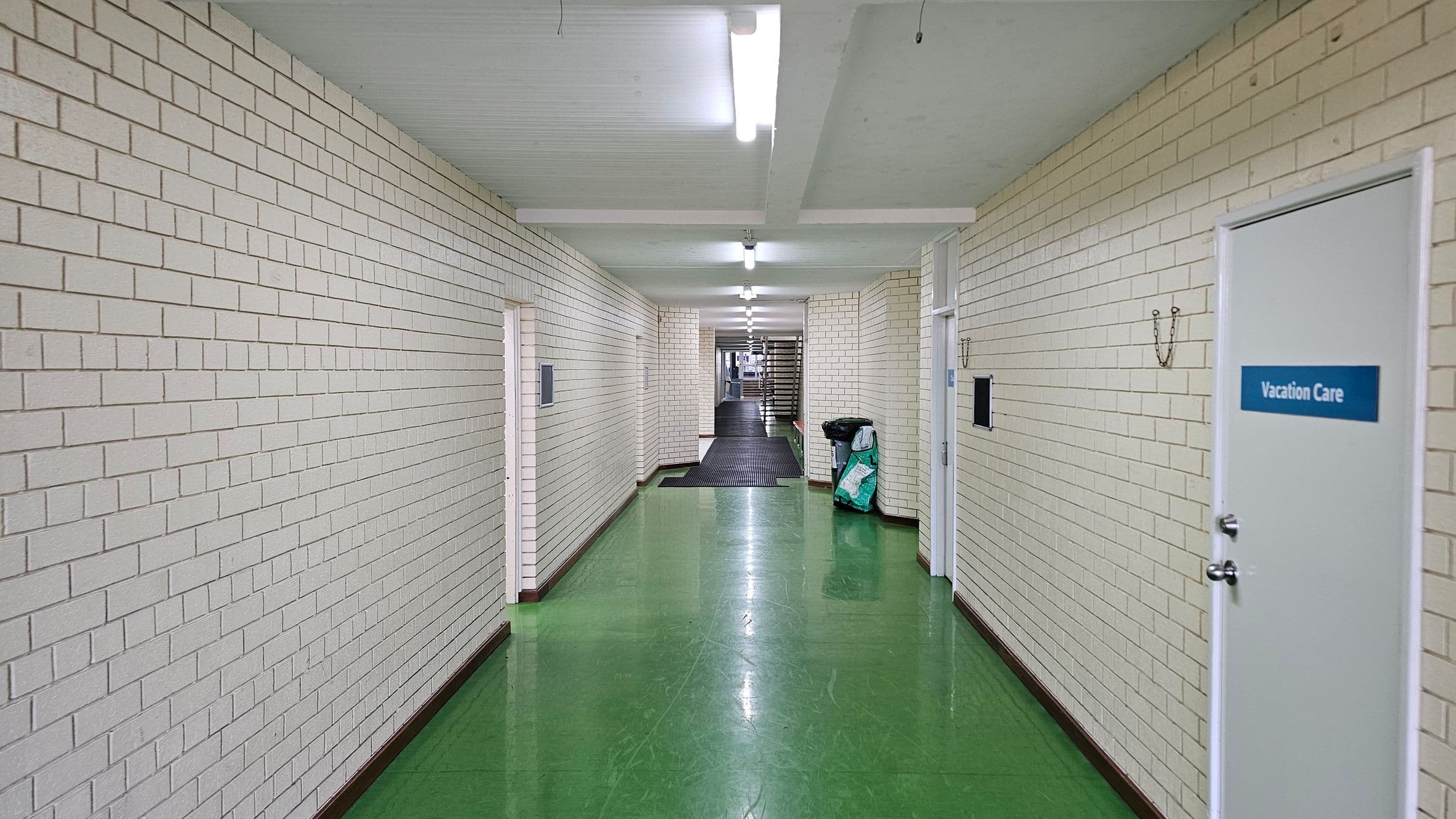 A long corridor with white brick walls and a shiny green floor. On the right, there is a door labelled "Vacation Care" and a black bin with a bag beside it. The ceiling is white with fluorescent lights evenly spaced along the length of the corridor. In the distance, the corridor narrows and leads to a staircase. The walls have hooks and small black panels mounted on them.
