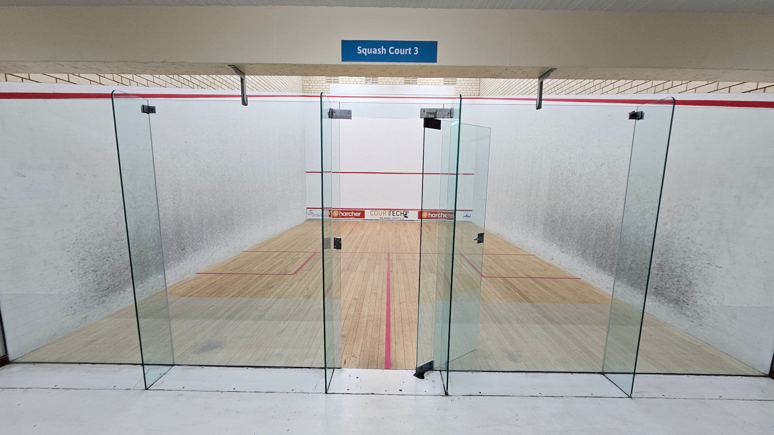 A squash court with a glass door entrance labelled "Squash Court 3" at the top. The court has wooden flooring with red boundary lines. The walls are white with a red line running horizontally near the top. Advertisements are visible at the far end of the court. The ceiling is light-coloured with visible beams.