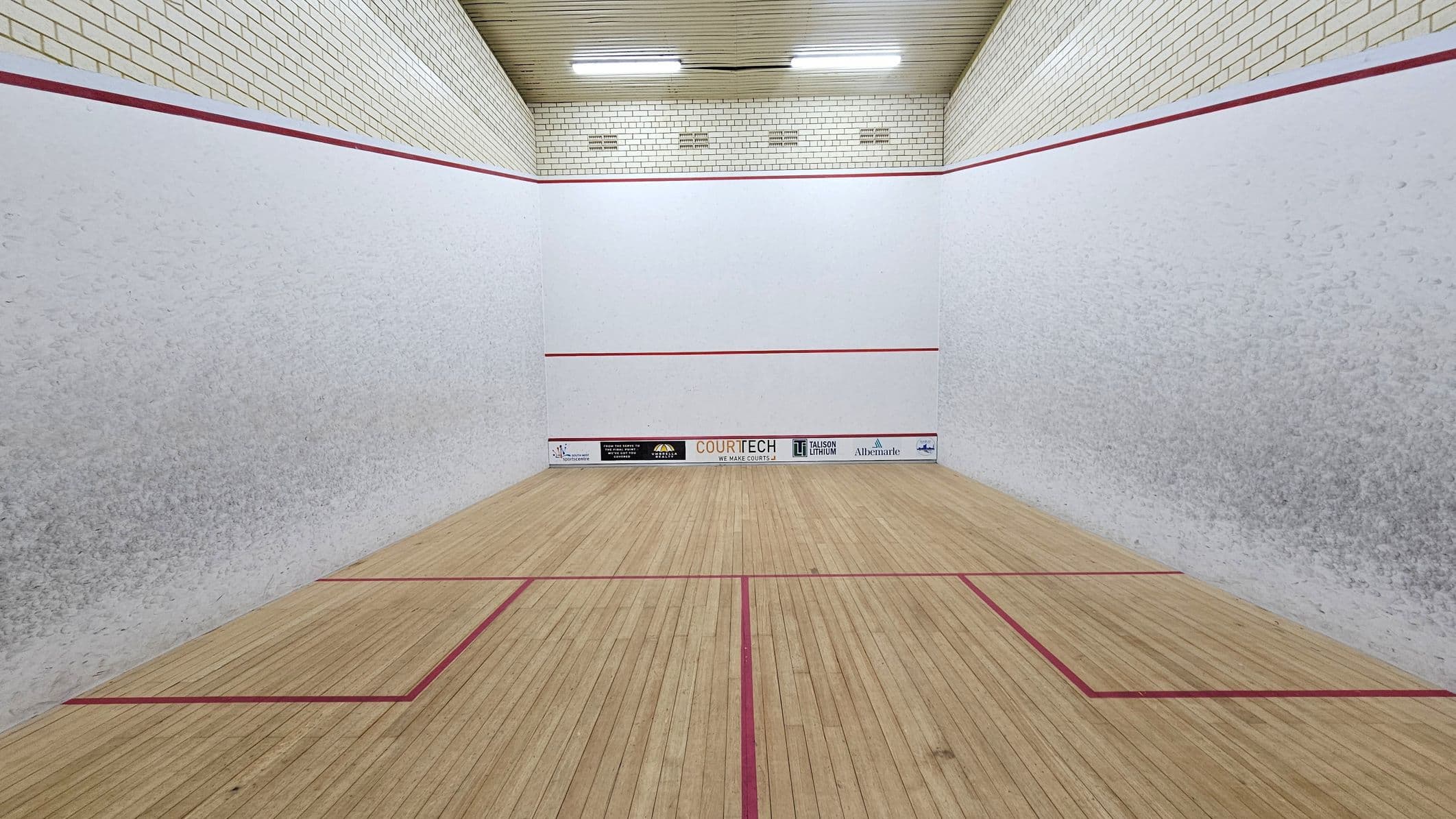A squash court with light wooden flooring marked by red lines. The walls are white with a red boundary line near the top. The back wall features several sponsor logos at the bottom. The ceiling is wooden with fluorescent lights. The side walls have a slightly textured appearance.