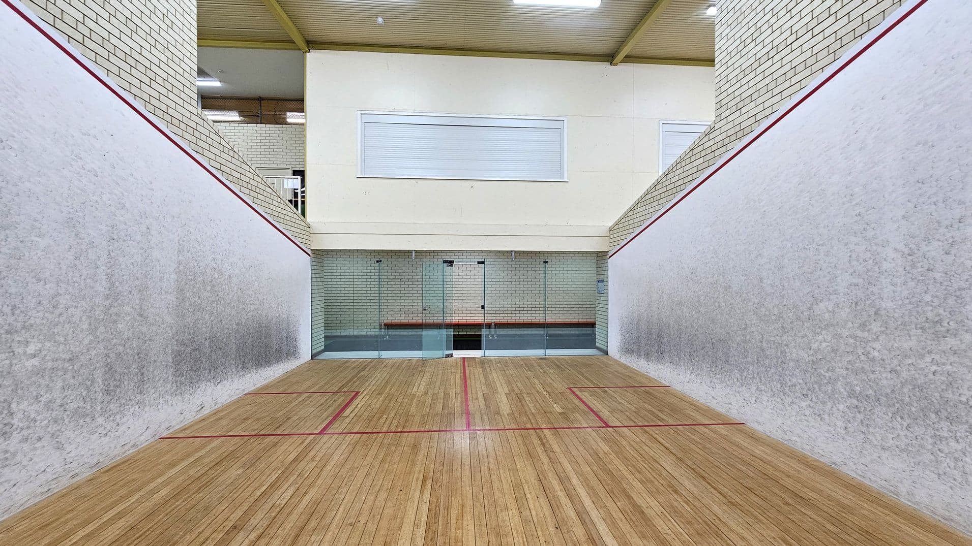A squash court with wooden flooring marked by red lines. The walls are light grey with a red outline near the top. The back wall is made of glass panels, allowing a view into an adjacent area with white brick walls. Above the glass, there is a white panel with a closed window. The ceiling is wooden with visible beams and fluorescent lights.