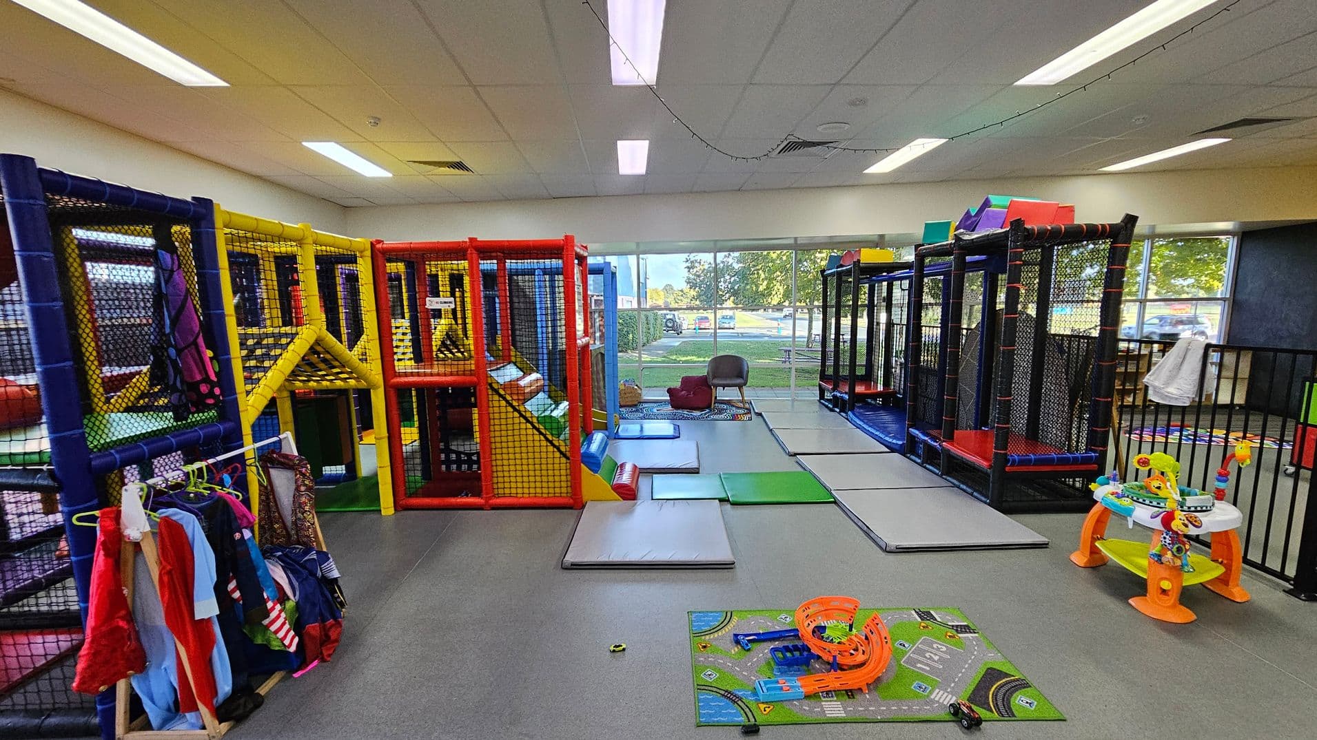 Indoor play area with colourful soft play structures on the left, featuring yellow, red, and blue frames with netting. A rack of children's costumes is nearby. In the centre, padded mats are spread on the floor. On the right, a black-framed play structure and a small activity table with toys are visible. Large windows in the background show an outdoor view with trees and parked cars. The ceiling has fluorescent lights and a grey floor.
