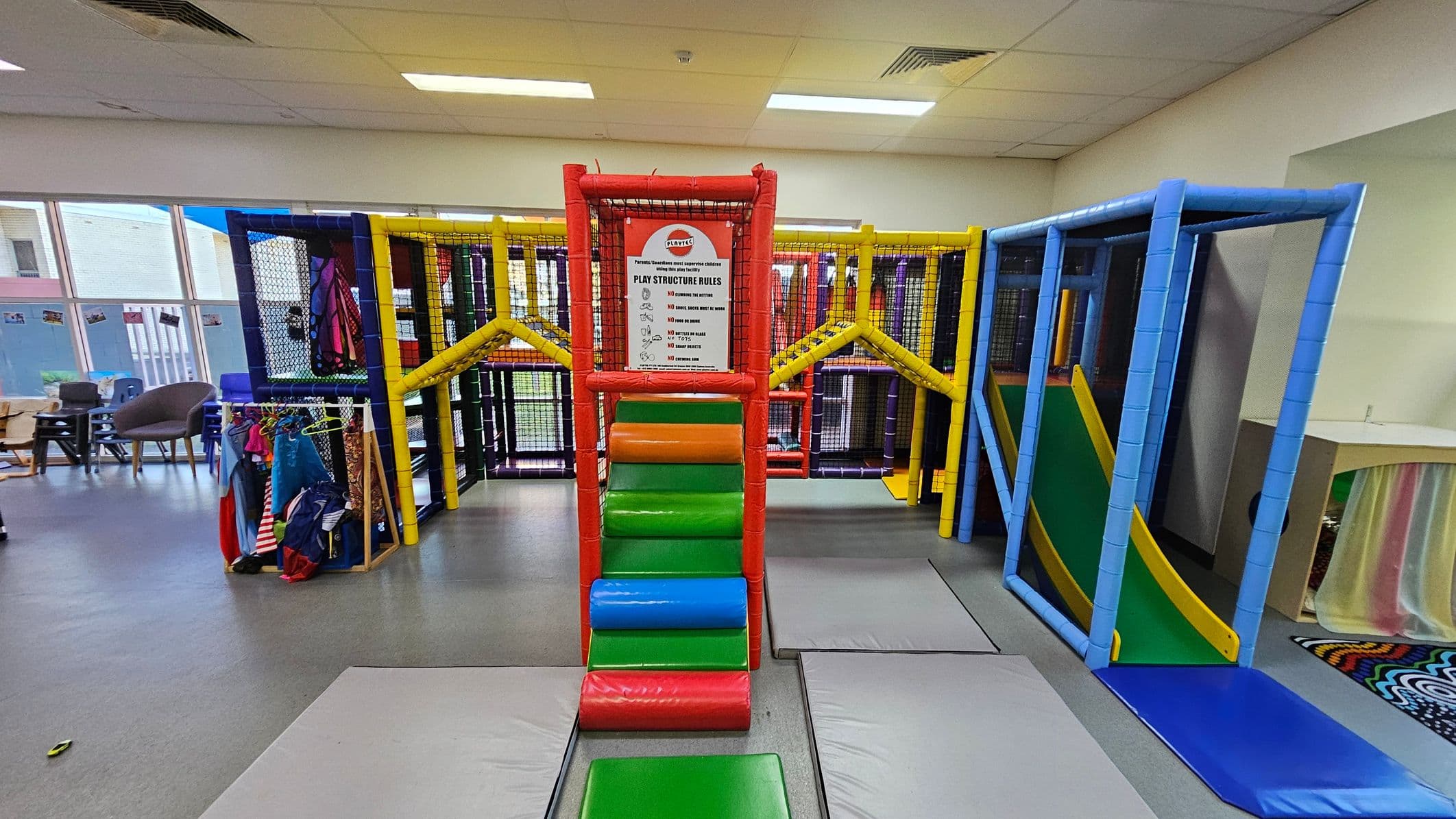 Indoor play area with a colourful climbing structure in the centre, featuring red, green, orange, and blue padded steps leading up to a platform. A sign with play structure rules is displayed at the top. On the left, there are chairs and a rack with children's costumes. On the right, a slide with a yellow border and green surface is visible. The floor is grey with padded mats, and the ceiling has fluorescent lights. Large windows are in the background.