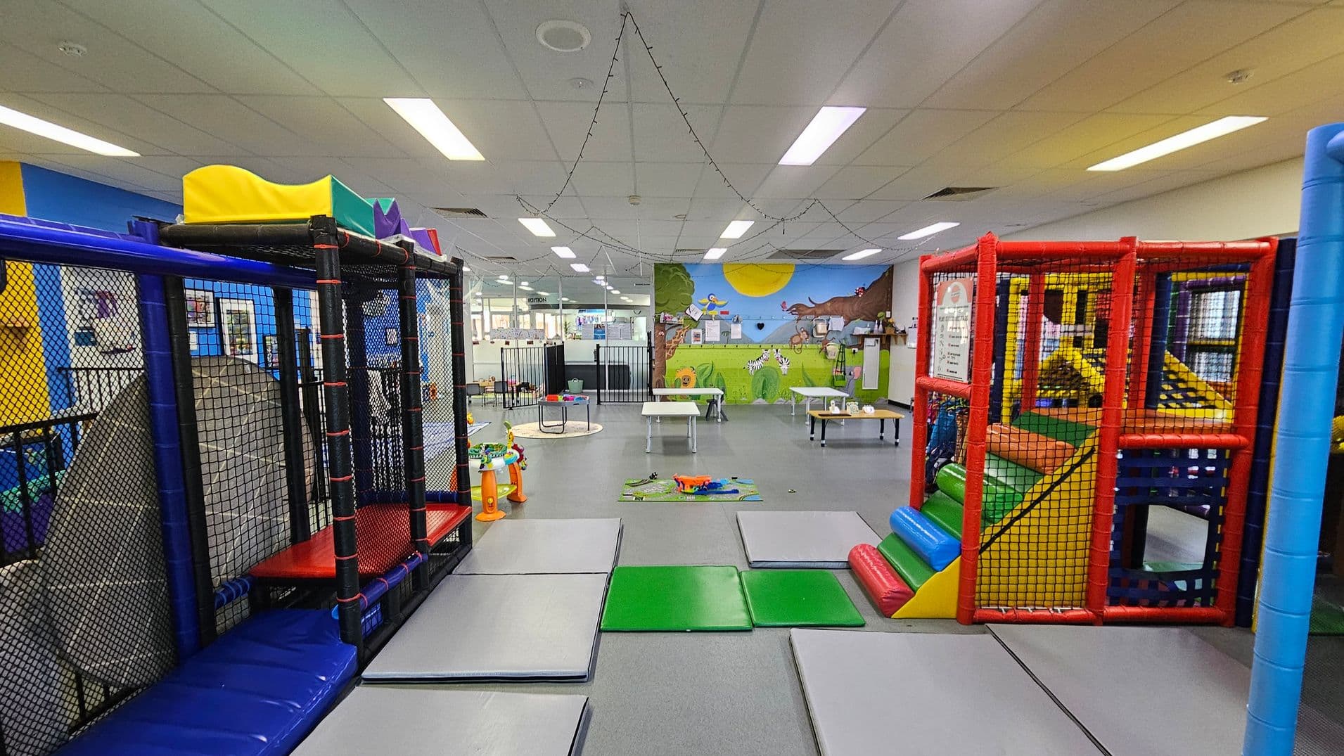 A colourful indoor play area with padded climbing structures on the left and right, featuring netted enclosures and slides. The floor is covered with grey and green mats. In the middle, there are small tables and toys scattered around. The back wall has a vibrant mural depicting a sunny outdoor scene with animals. The ceiling is lined with fluorescent lights and string lights.