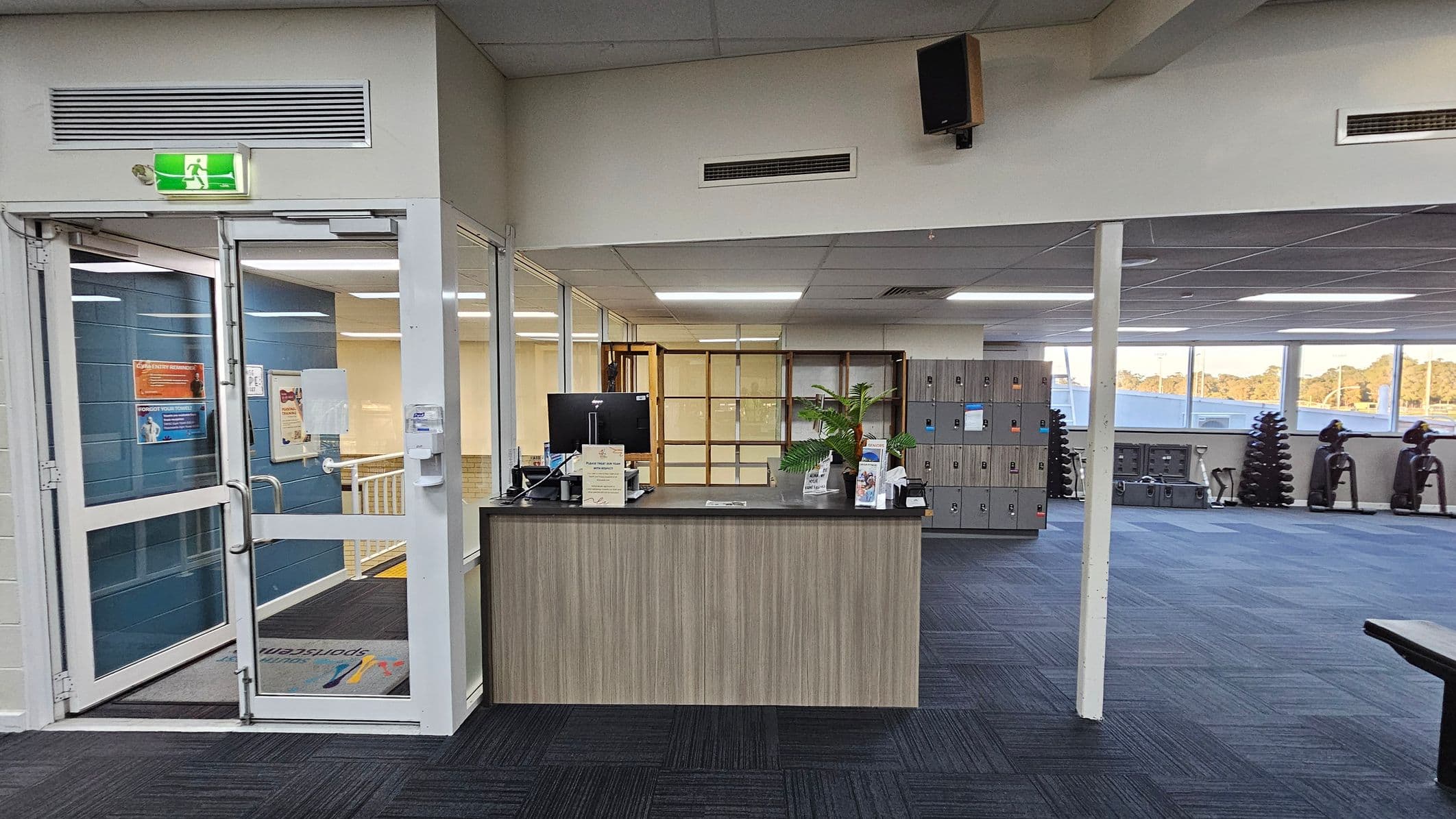 A reception area with a wooden desk in the centre, featuring a computer monitor, brochures, and a potted plant. To the left, a glass door with an illuminated exit sign above it leads to a hallway. On the right, a row of lockers is visible, along with exercise equipment like stationary bikes and stacked weights. The floor is carpeted in dark and light blue squares, and the ceiling has fluorescent lighting. Large windows in the background let in natural light.