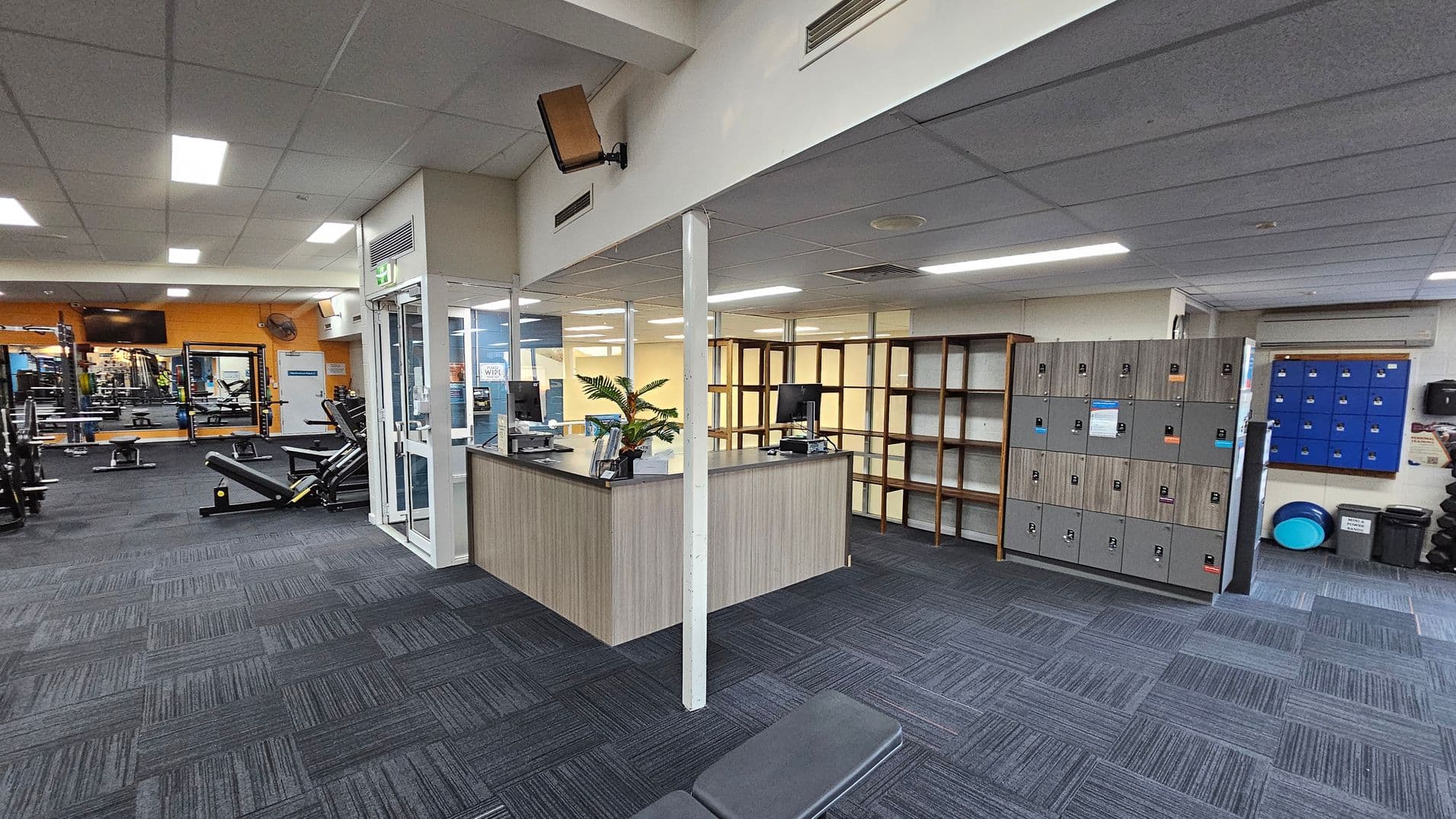 A gym interior with a reception desk in the centre, featuring a computer and a plant. To the right, there are grey lockers and blue lockers on the wall. The left side has gym equipment, including benches and weight racks. The floor is covered with patterned carpet tiles in shades of grey and purple. The ceiling is fitted with fluorescent lights.