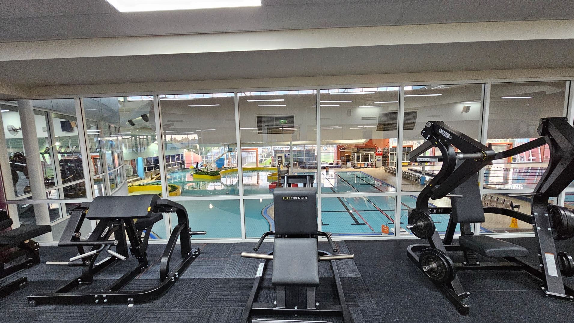 In the foreground, there are various gym machines on a dark carpeted floor. The middle features a large glass wall separating the gym area from an indoor swimming pool. The pool area has lanes and a yellow water slide. The ceiling is white with fluorescent lights. The background shows more gym equipment and a colourful pool area with windows allowing natural light.